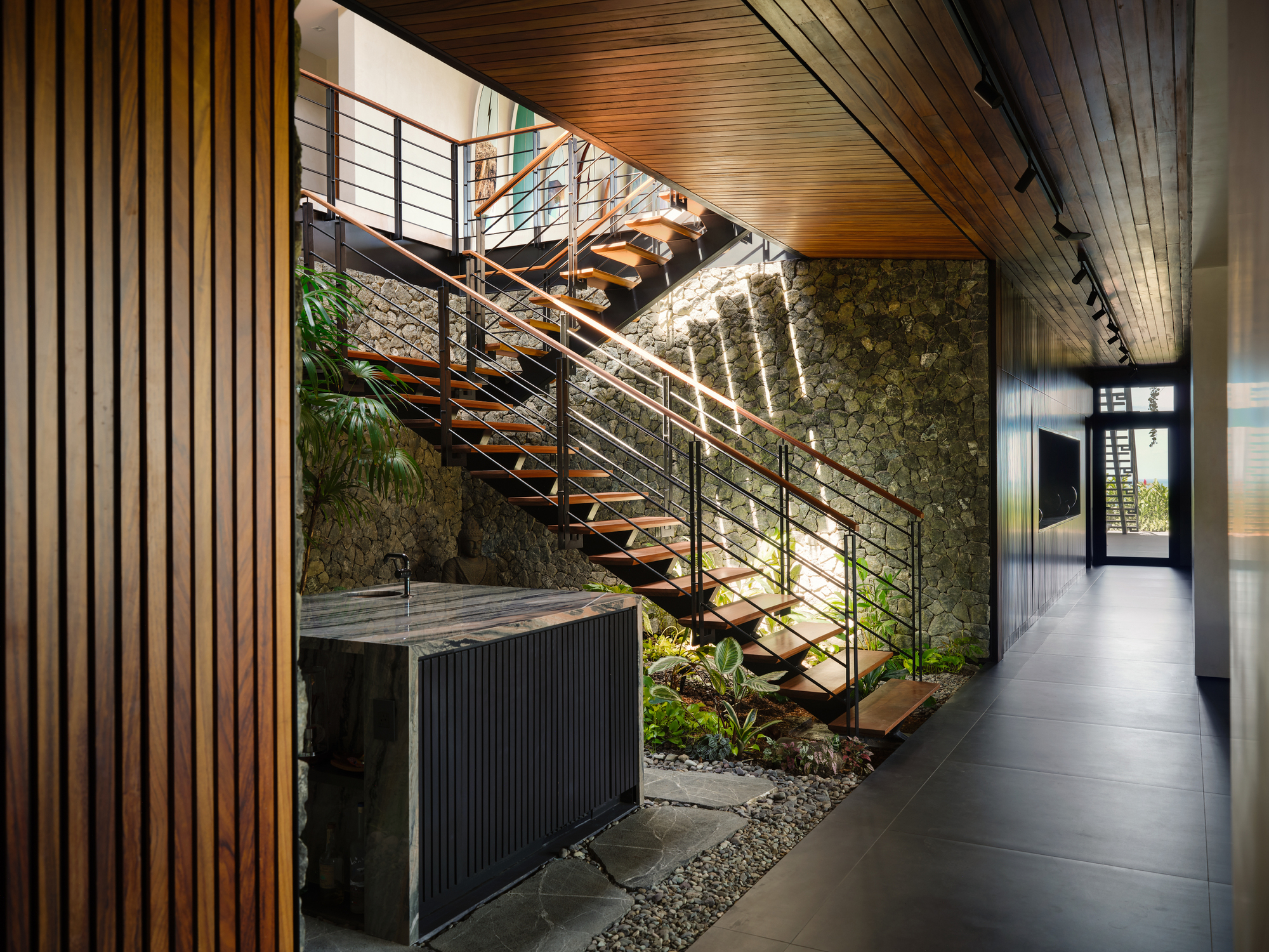 Open-tread timber staircase rising beside a rough stone wall, with a stone sink and pebble planting bed below
