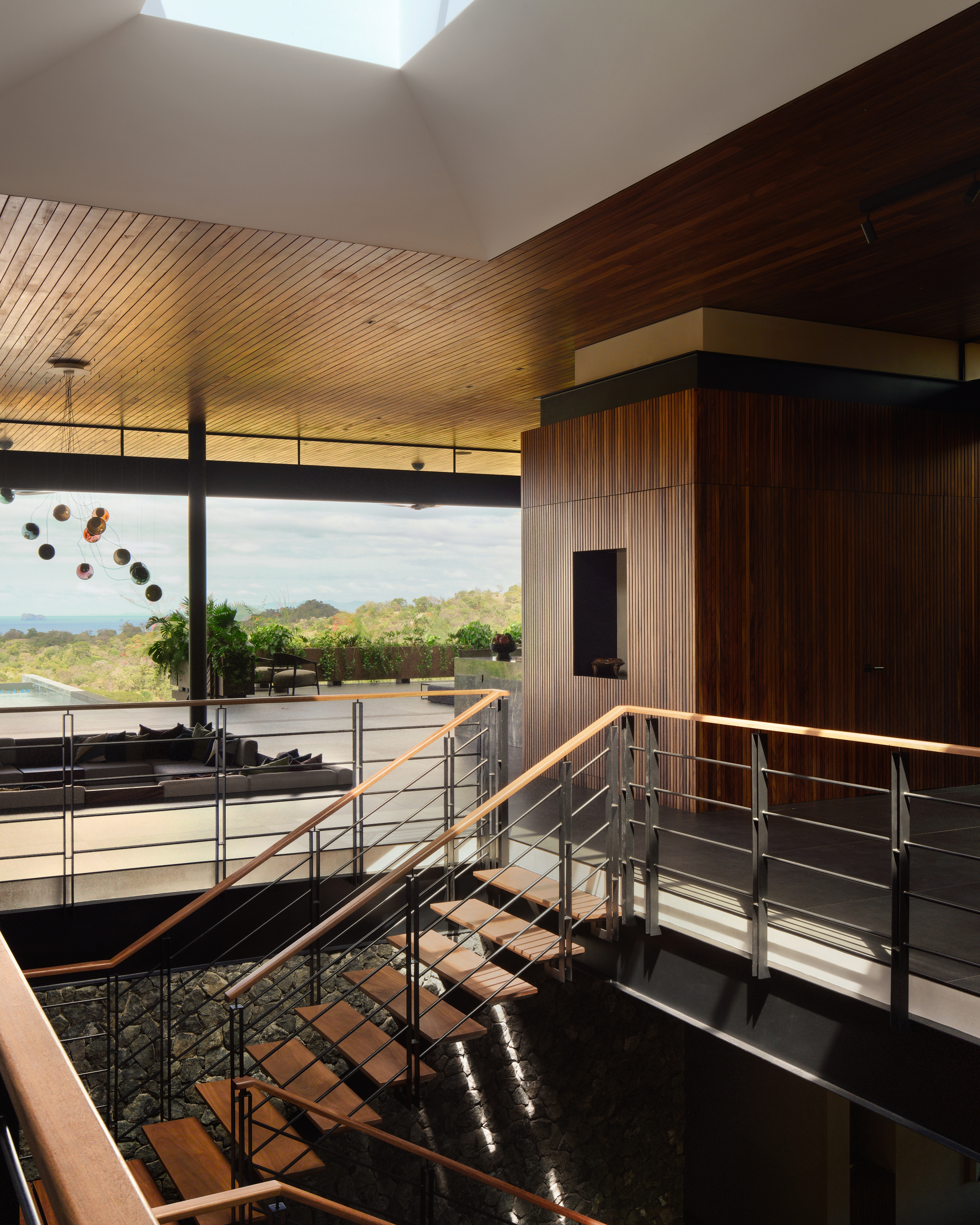 View from the stair landing across a stone-lined void toward the terrace, with hills and distant sea under cloud