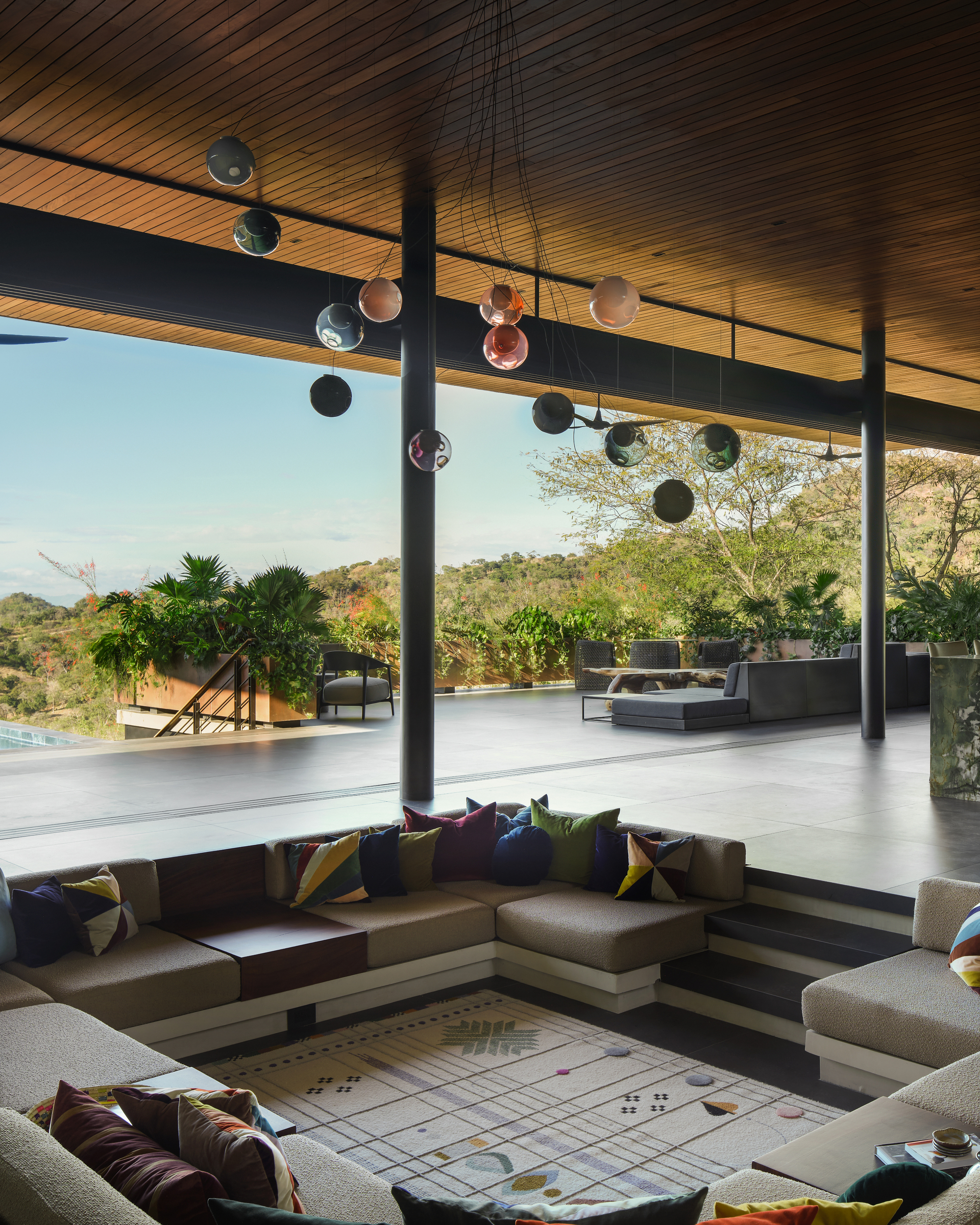 Sunken lounge with cushioned seating and patterned rug beneath a timber ceiling, glass spheres hanging above and green hills beyond