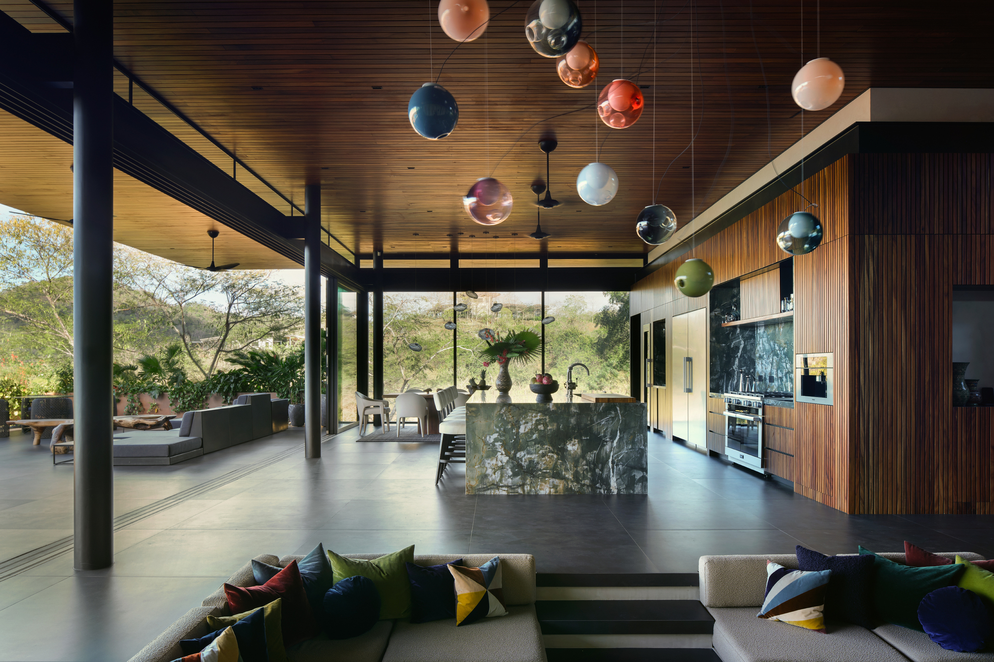 Open living area with a marble-clad island and striped timber kitchen wall, colorful pendants hanging from the ceiling in afternoon light