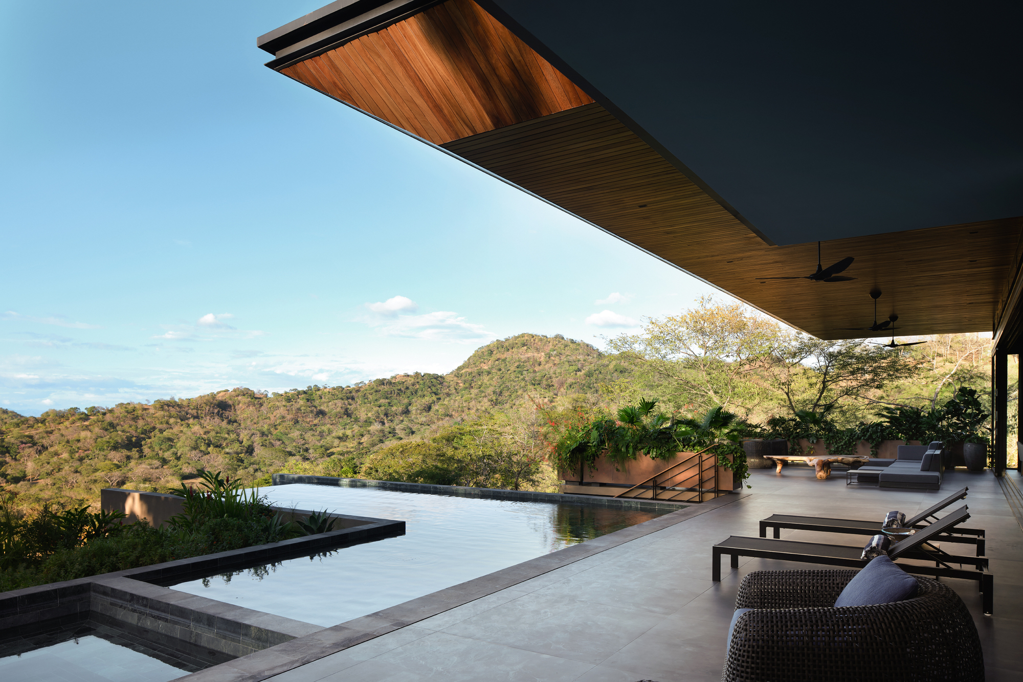 Infinity pool edge and cantilevered timber-lined roof over a terrace with loungers, facing forested hills under a clear blue sky