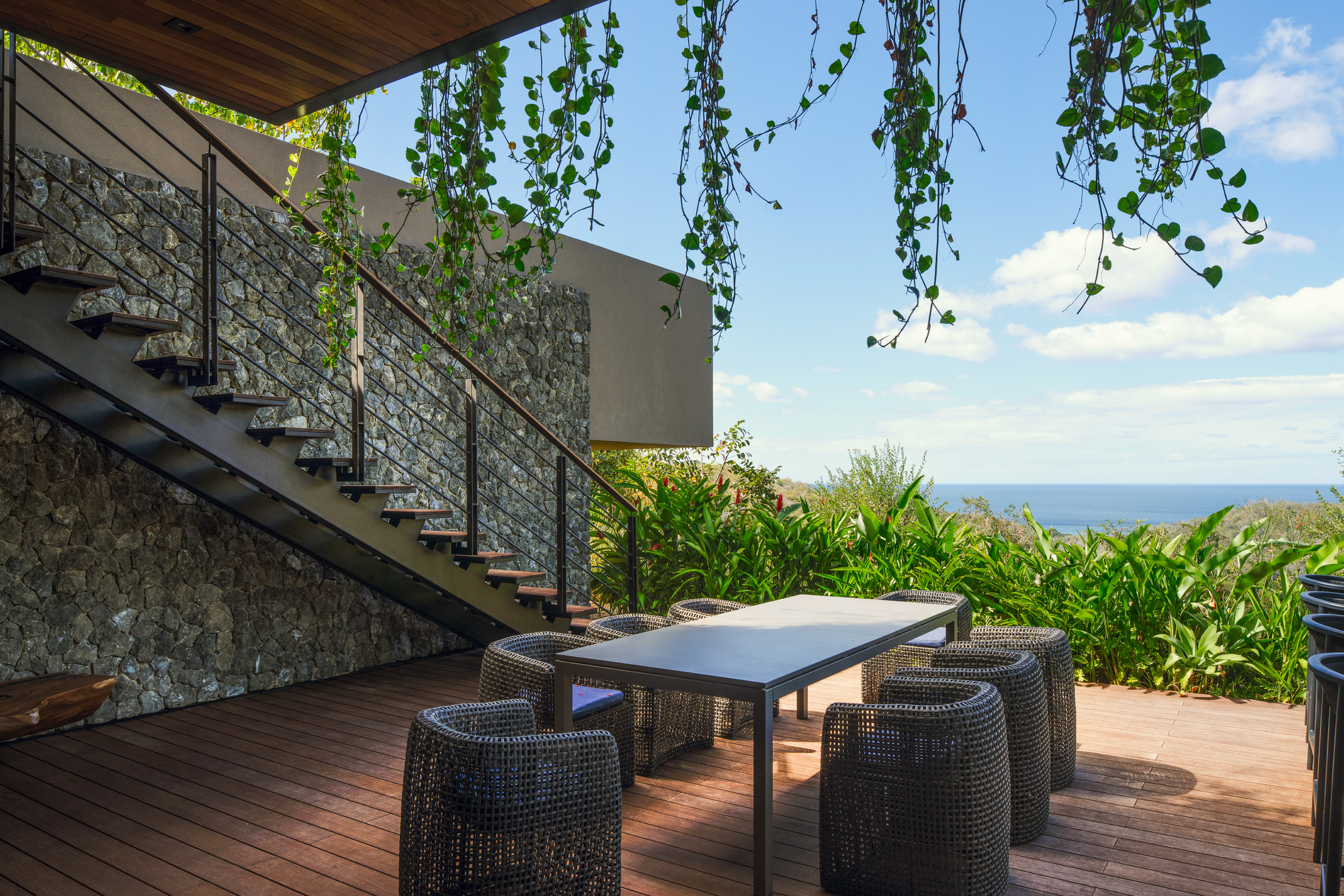 Dining table and woven chairs on a timber deck beside a stone wall and steel stair, with vines hanging overhead