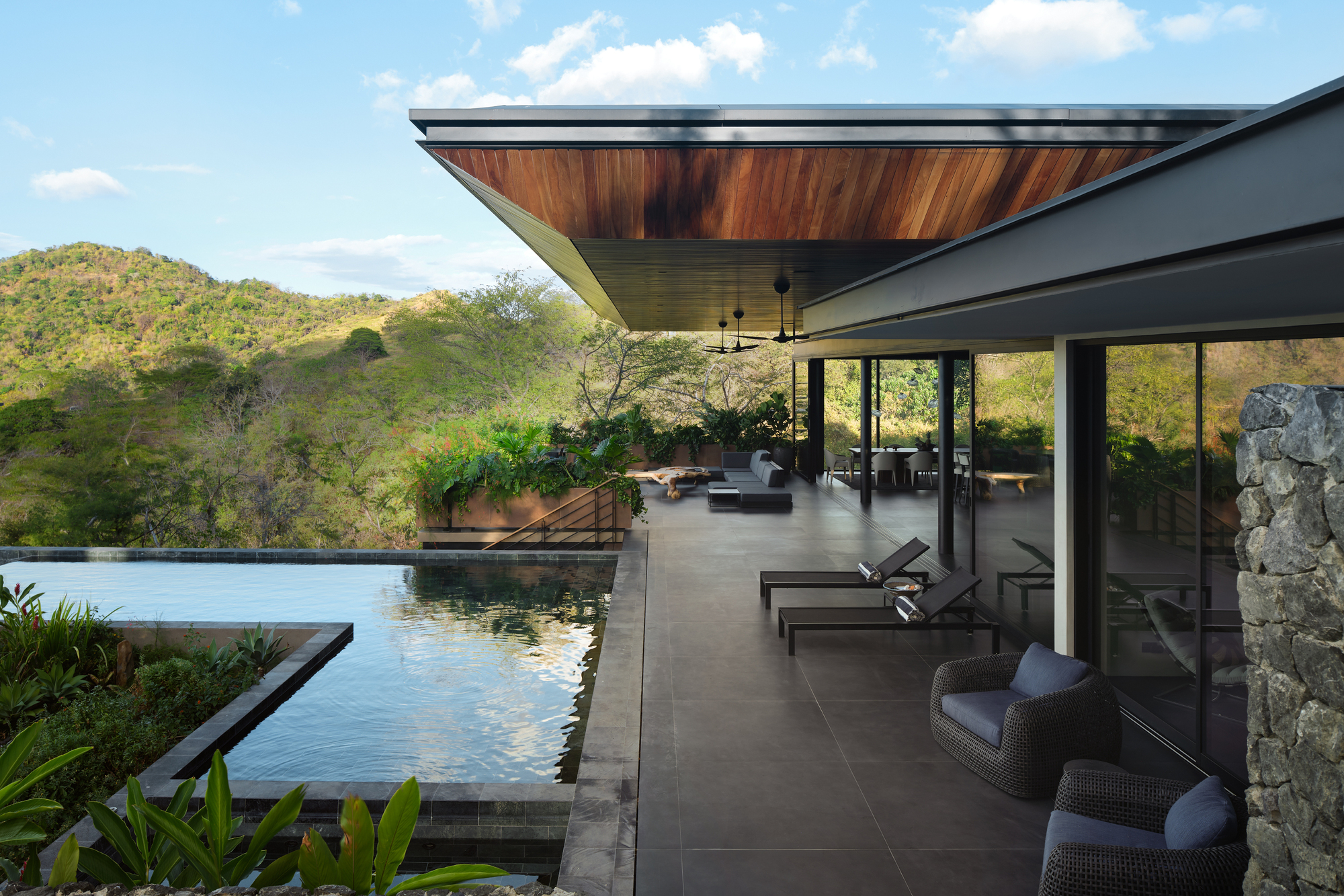 Terrace with loungers and wicker chairs beside the pool, a stone column in the foreground and green hills behind
