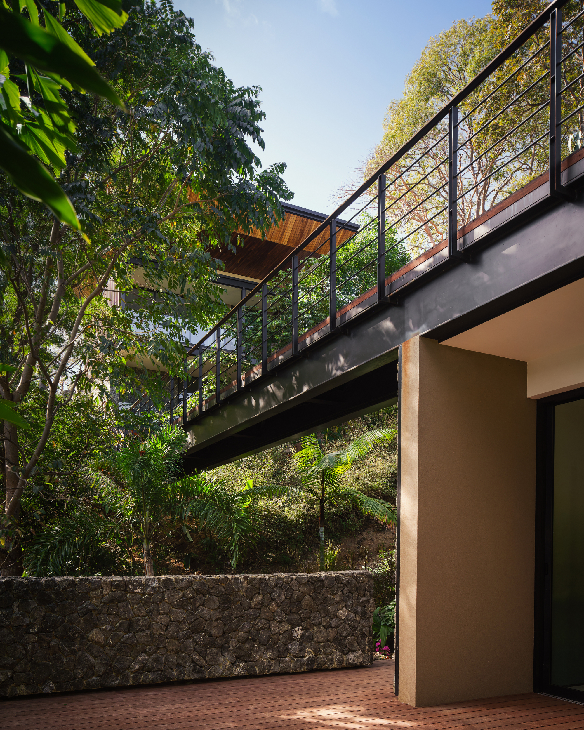 Steel-framed walkway with black railings spanning above a rough stone wall and timber deck, surrounded by hillside trees in sunlight