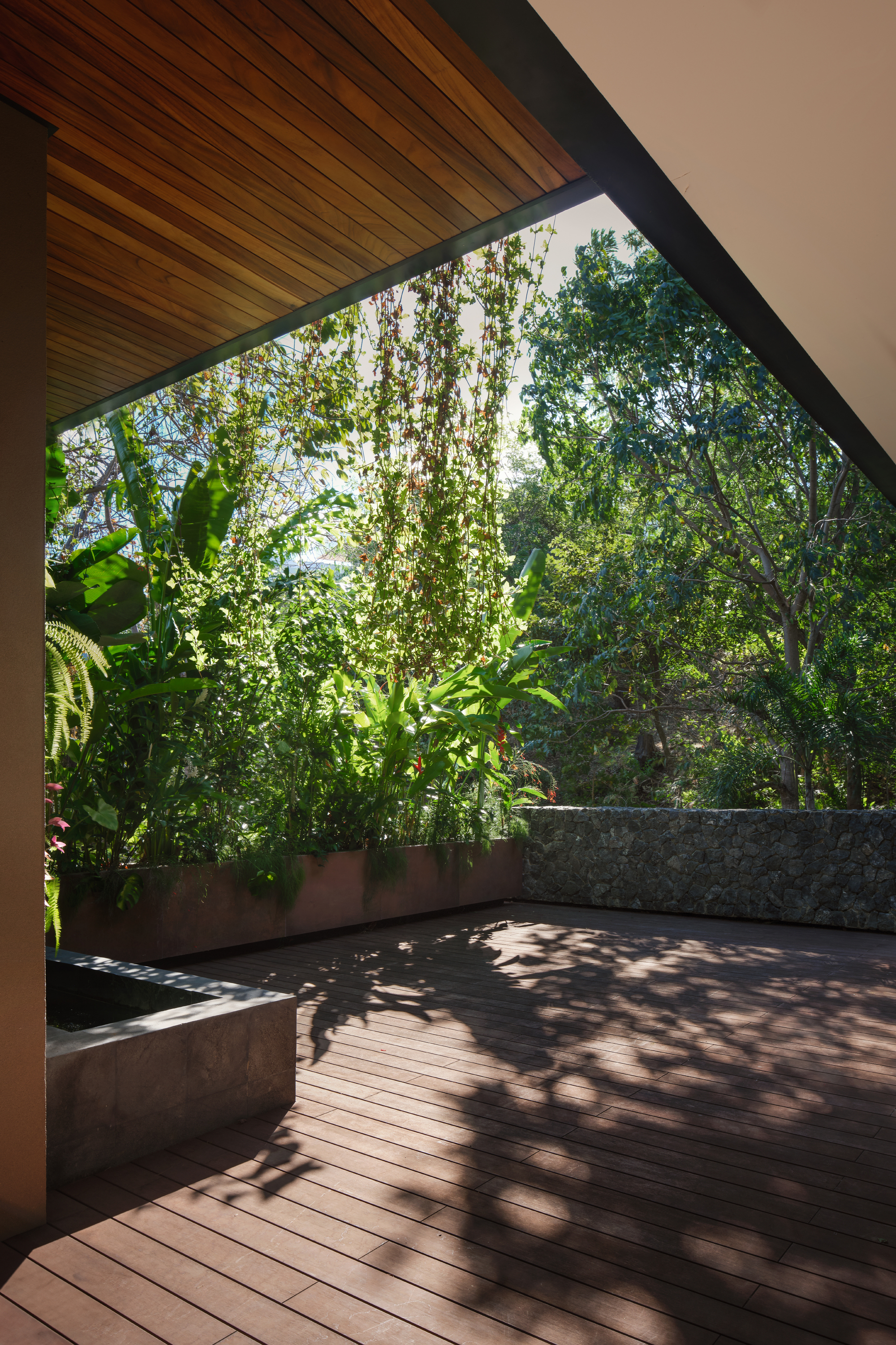 Covered timber deck beside a planted bed and stone wall, with dappled tree shadows falling across the boards