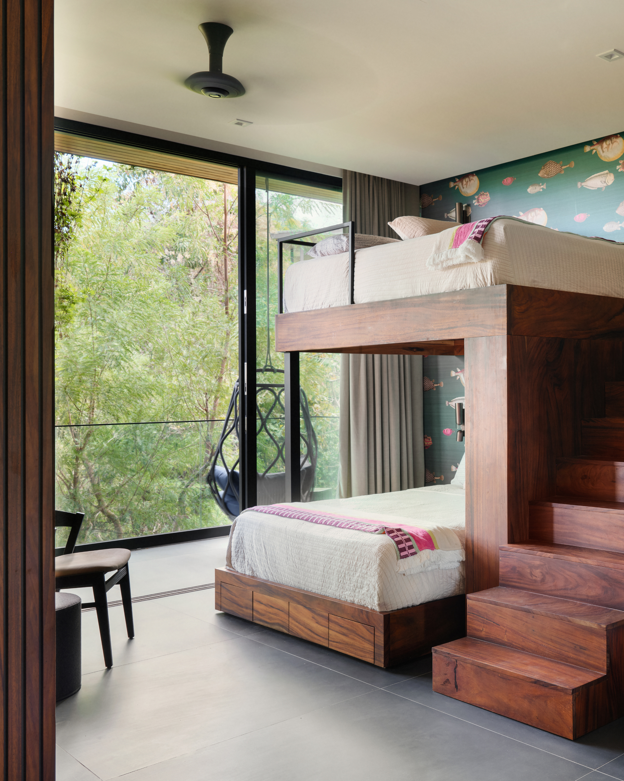 Wooden bunk bed with built-in stairs and drawers beside a sliding glass door and hanging chair, with fish wallpaper behind