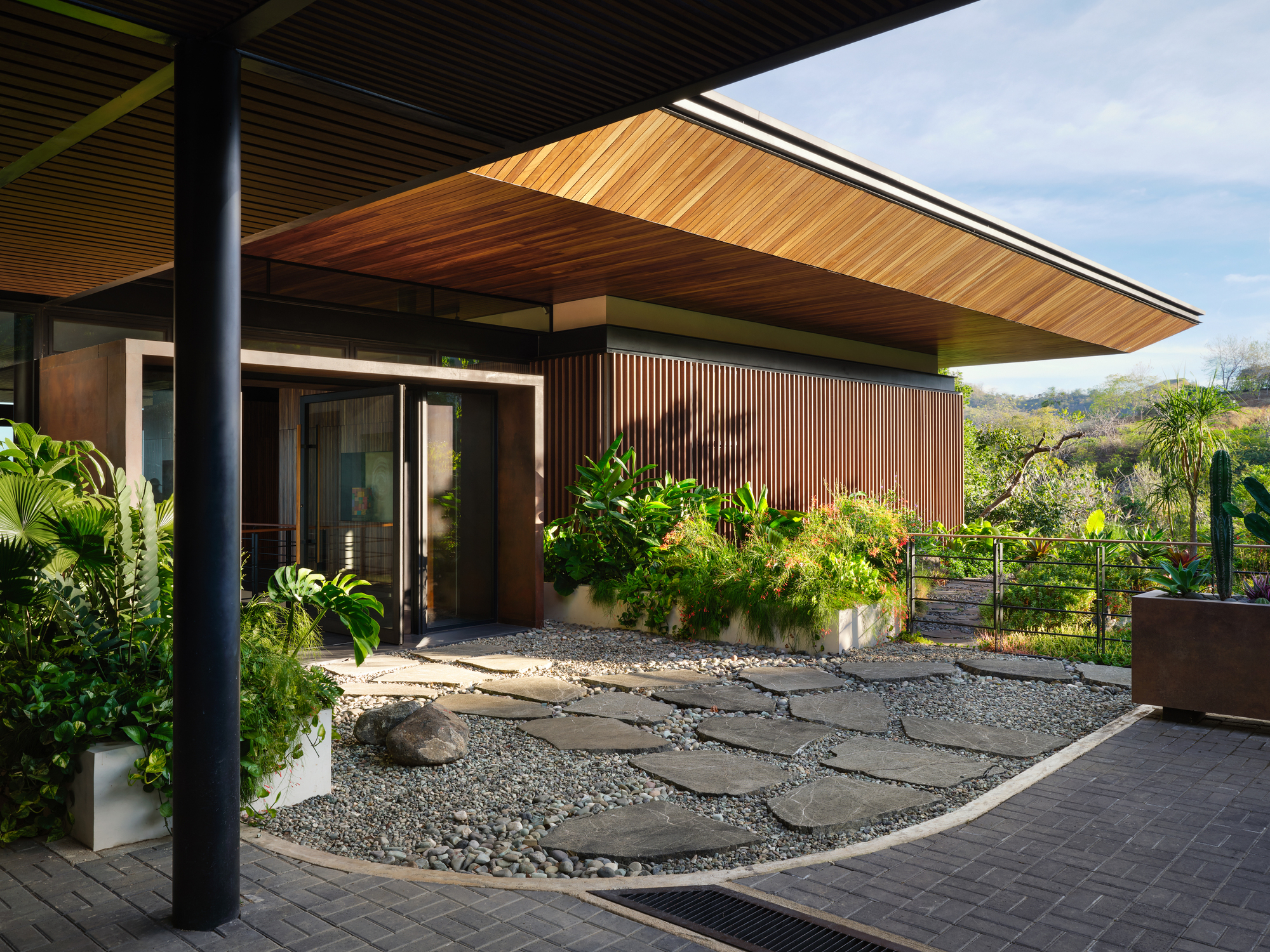 Entry courtyard with stepping stones across pebbles, planted beds and a cantilevered timber-clad roof under a pale afternoon sky