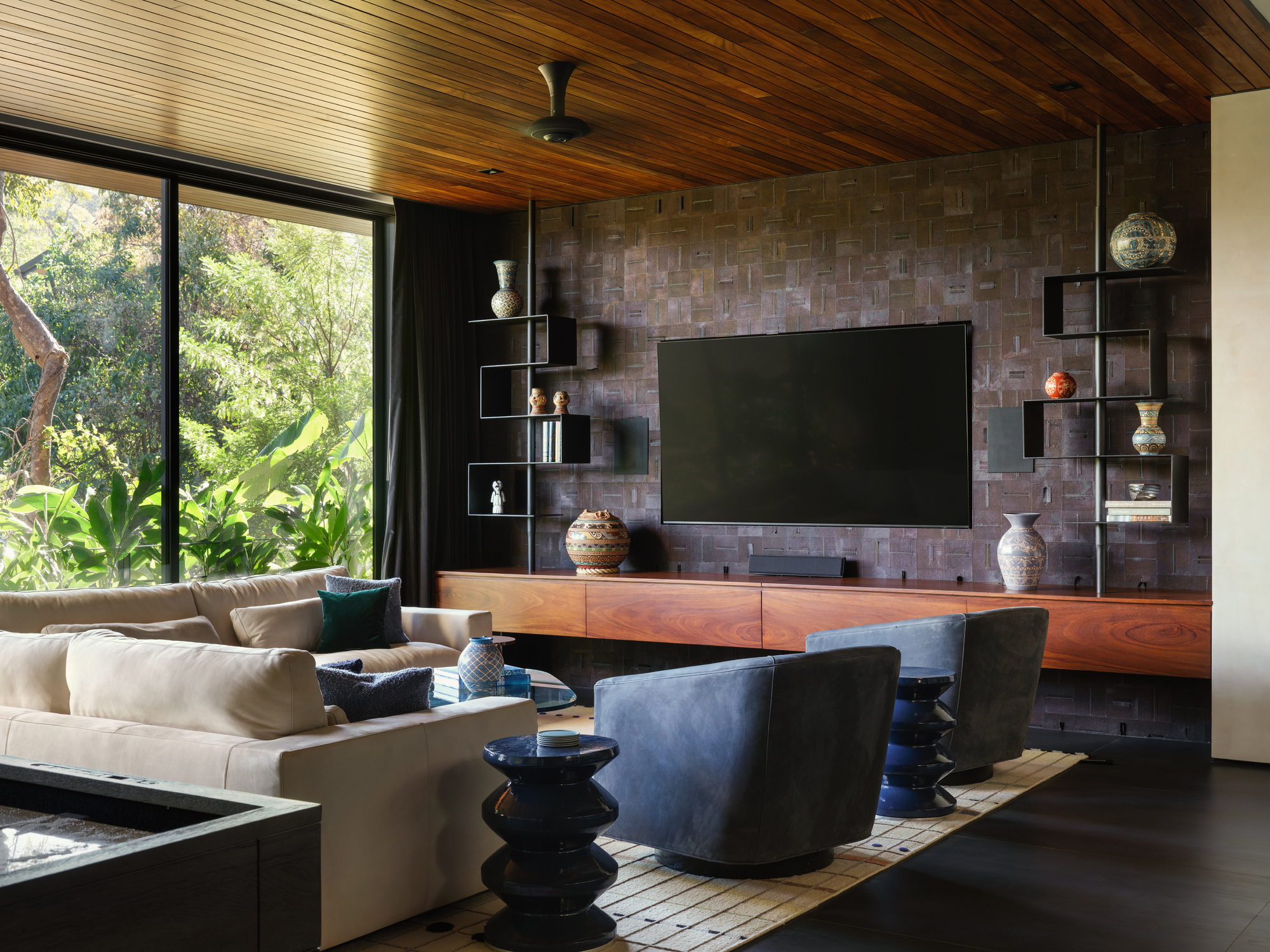 Living room with a woven-brick media wall, floating wood console and black shelving, beside a window onto green foliage