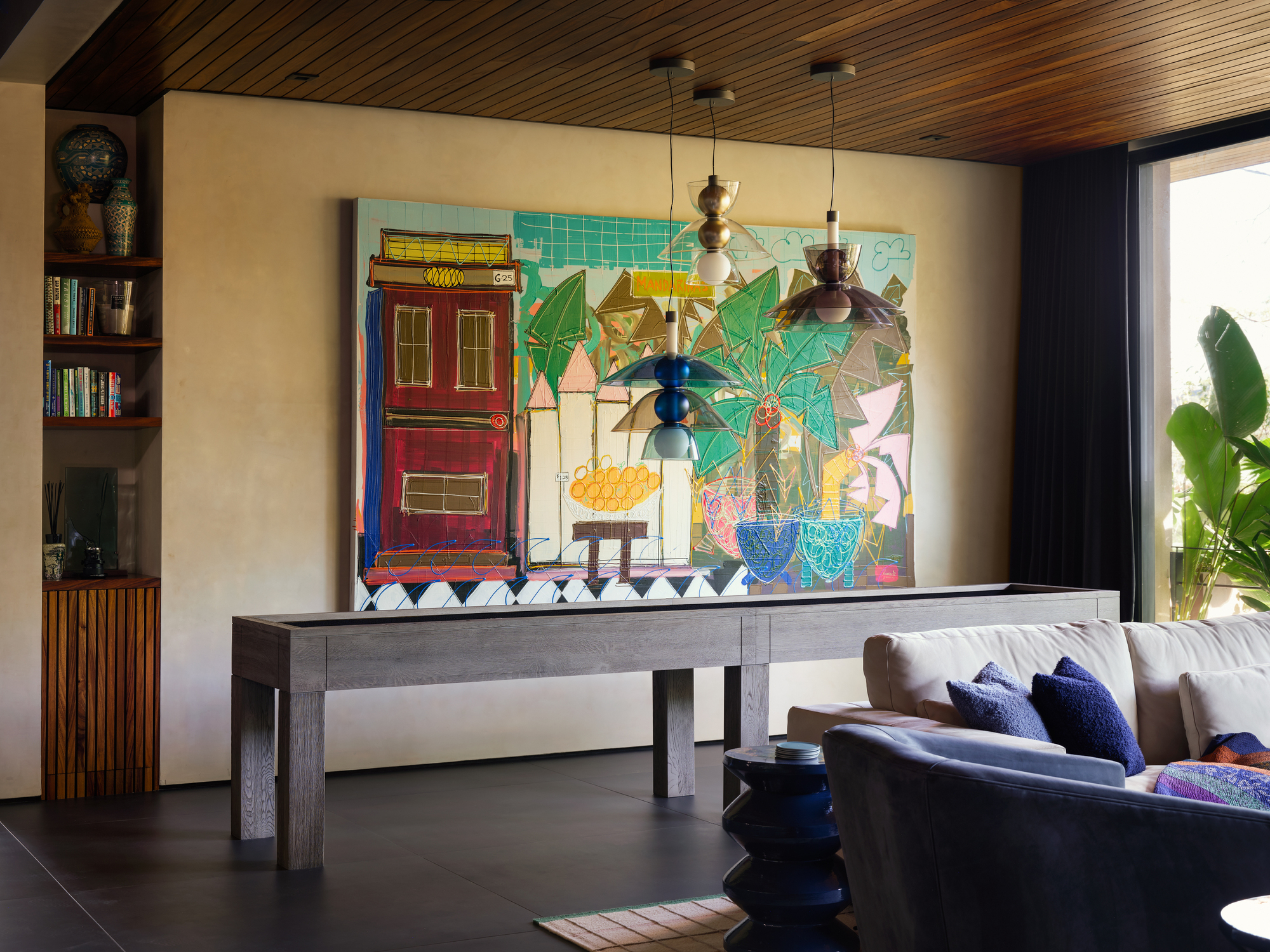 Colorful painted canvas above a long grey shuffleboard table, with three pendant lamps and a wood slat ceiling in warm daylight