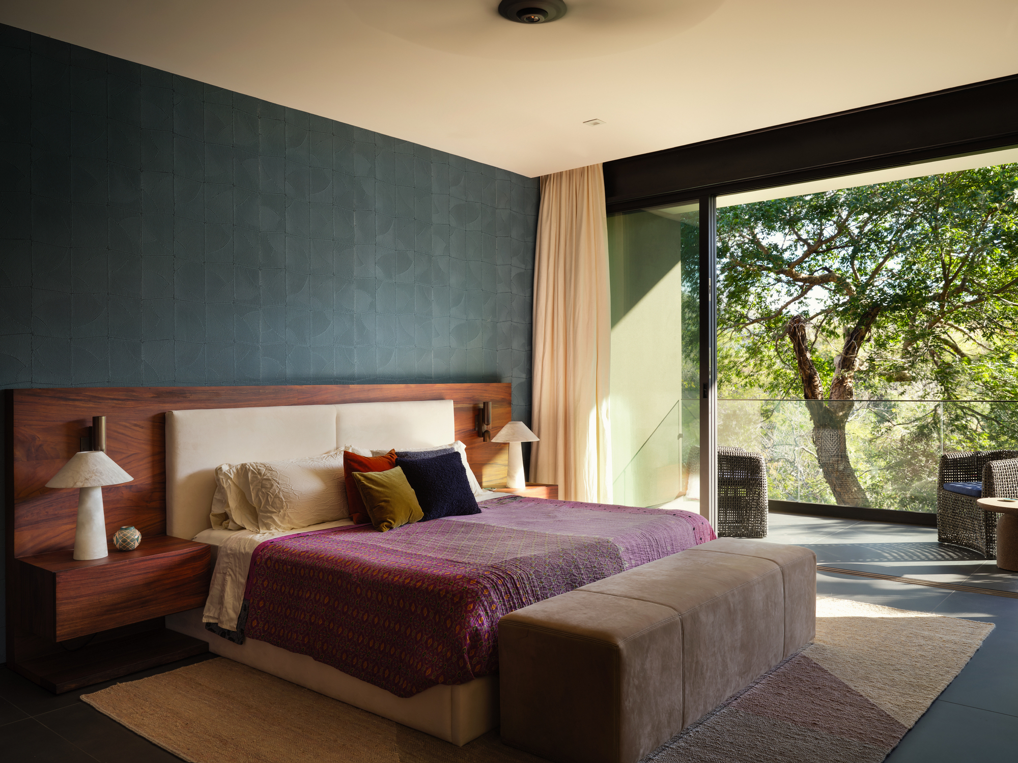 Bedroom with a walnut headboard, teal patterned wall and sliding glass door opening onto a tree in afternoon sun