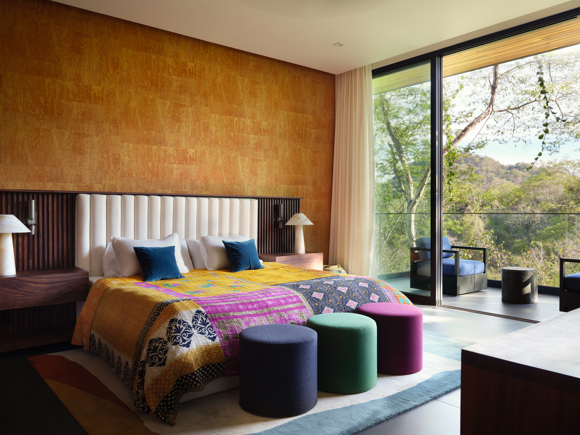 Bedroom with a gold textured wall, quilted bedspread and three round pouf stools beside a glass door facing forested hills