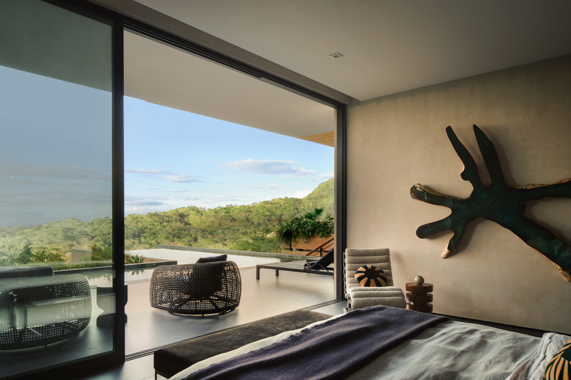 Bedroom view past the sliding glass door to a terrace with woven chairs and green hills under a pale sky