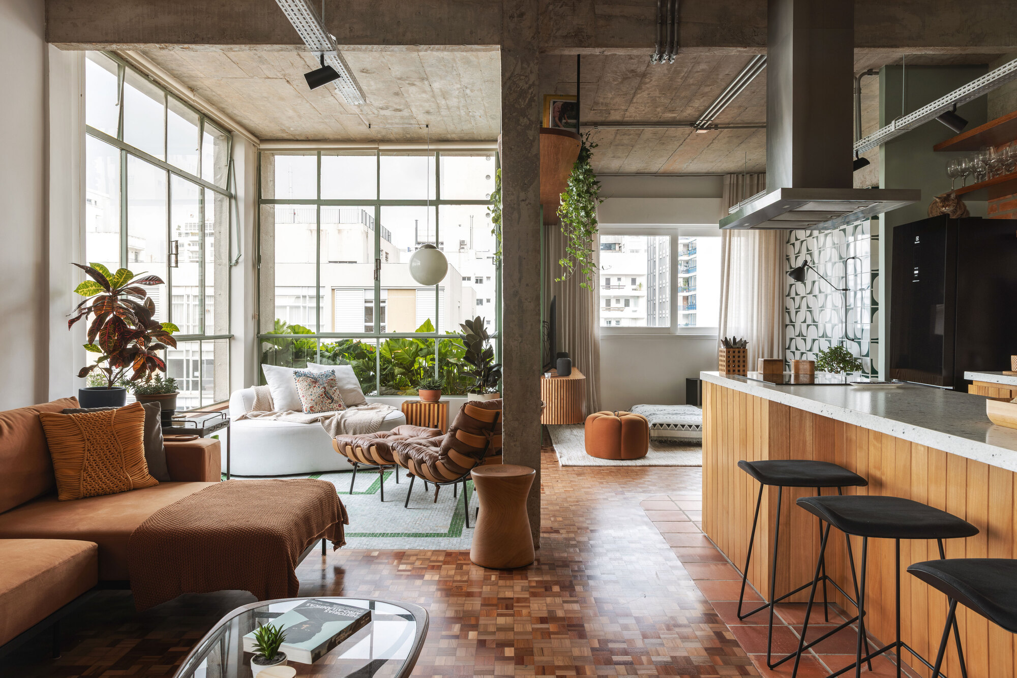 Open living area with a concrete column, parquet floor and timber-clad kitchen counter, with a corner of plants against tall steel-framed windows