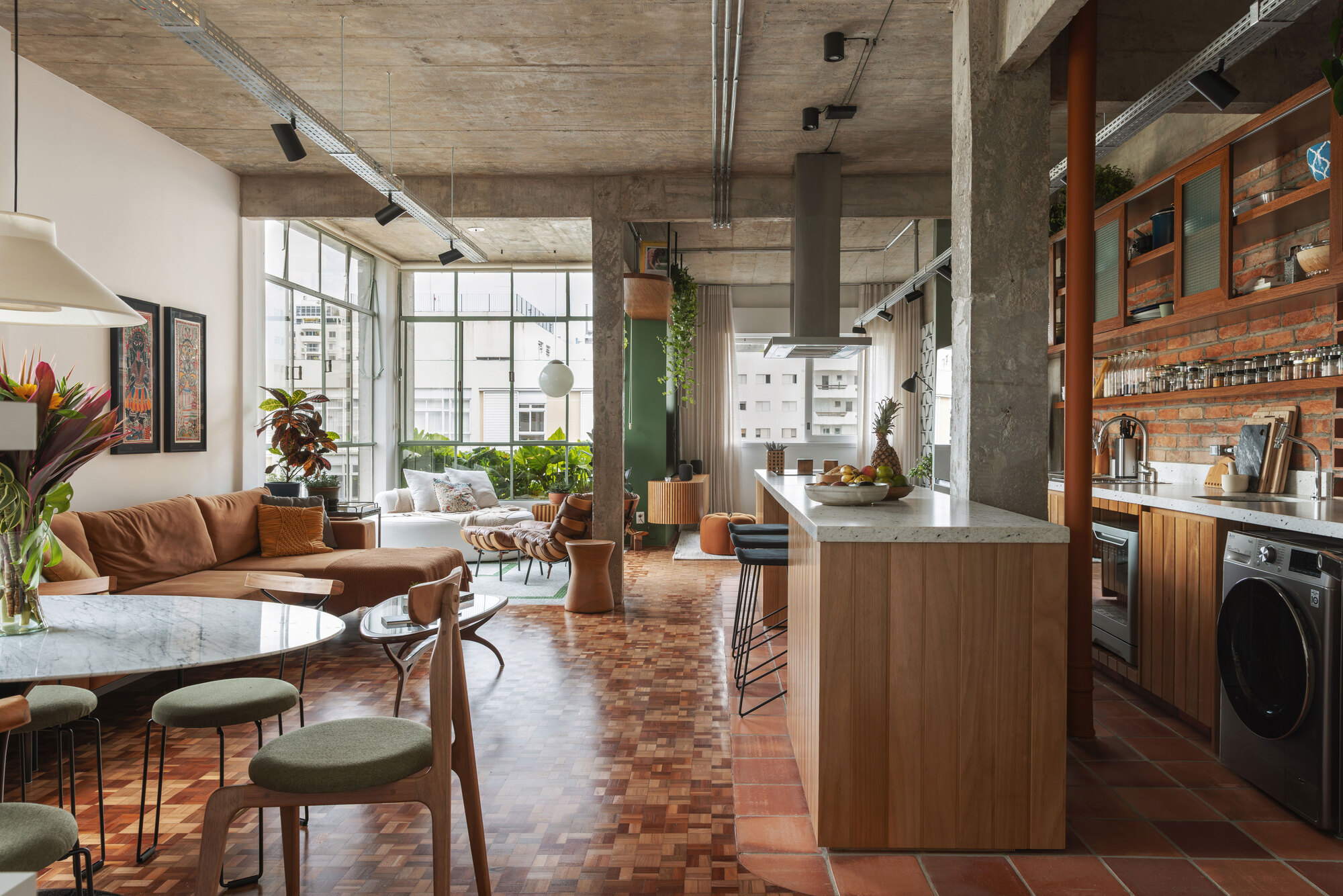 Open-plan view from dining table past an island counter to a brick-backed kitchen, with daylight from steel-framed windows beyond