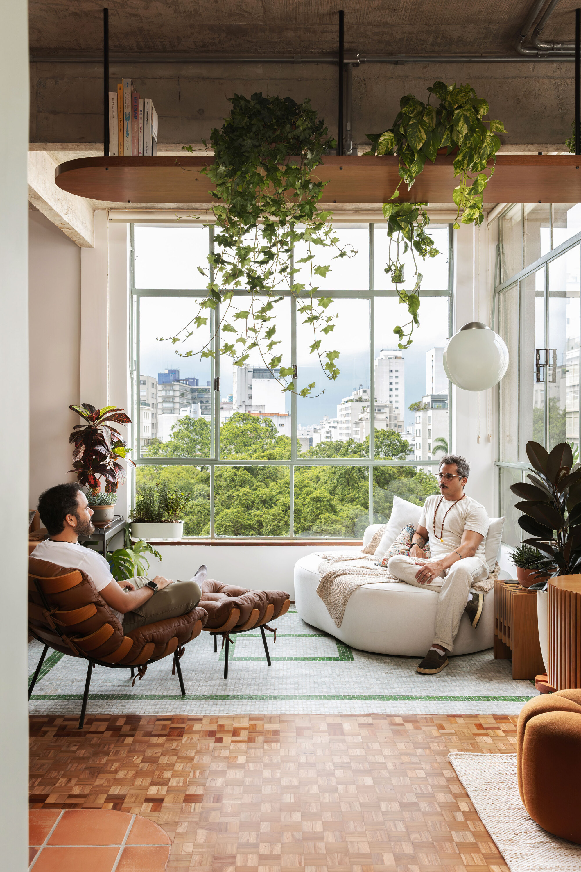 Two men talking in a window nook beneath trailing vines, with green tree canopy and distant towers outside