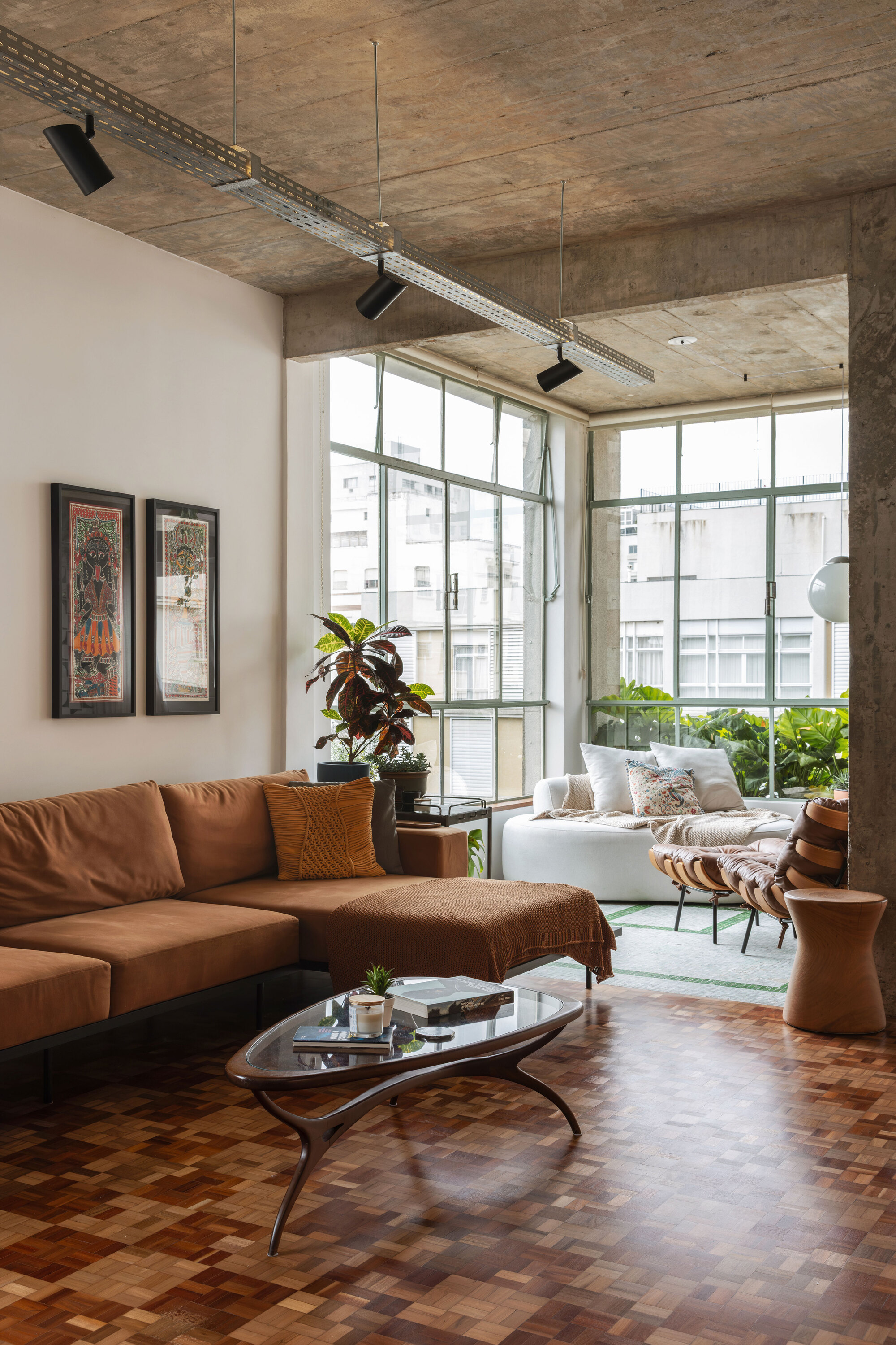 Corner of the living room with a rust-coloured sectional, oval glass coffee table and exposed concrete ceiling with cable tray lighting