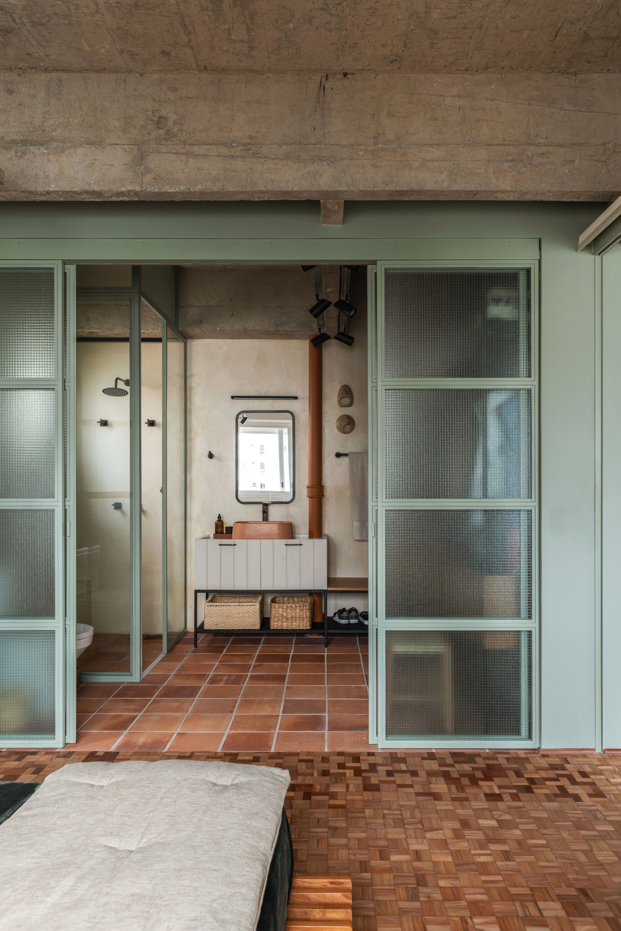 Bathroom seen through sage green framed sliding doors with reeded glass, terracotta floor tiles and a wood parquet floor in the foreground