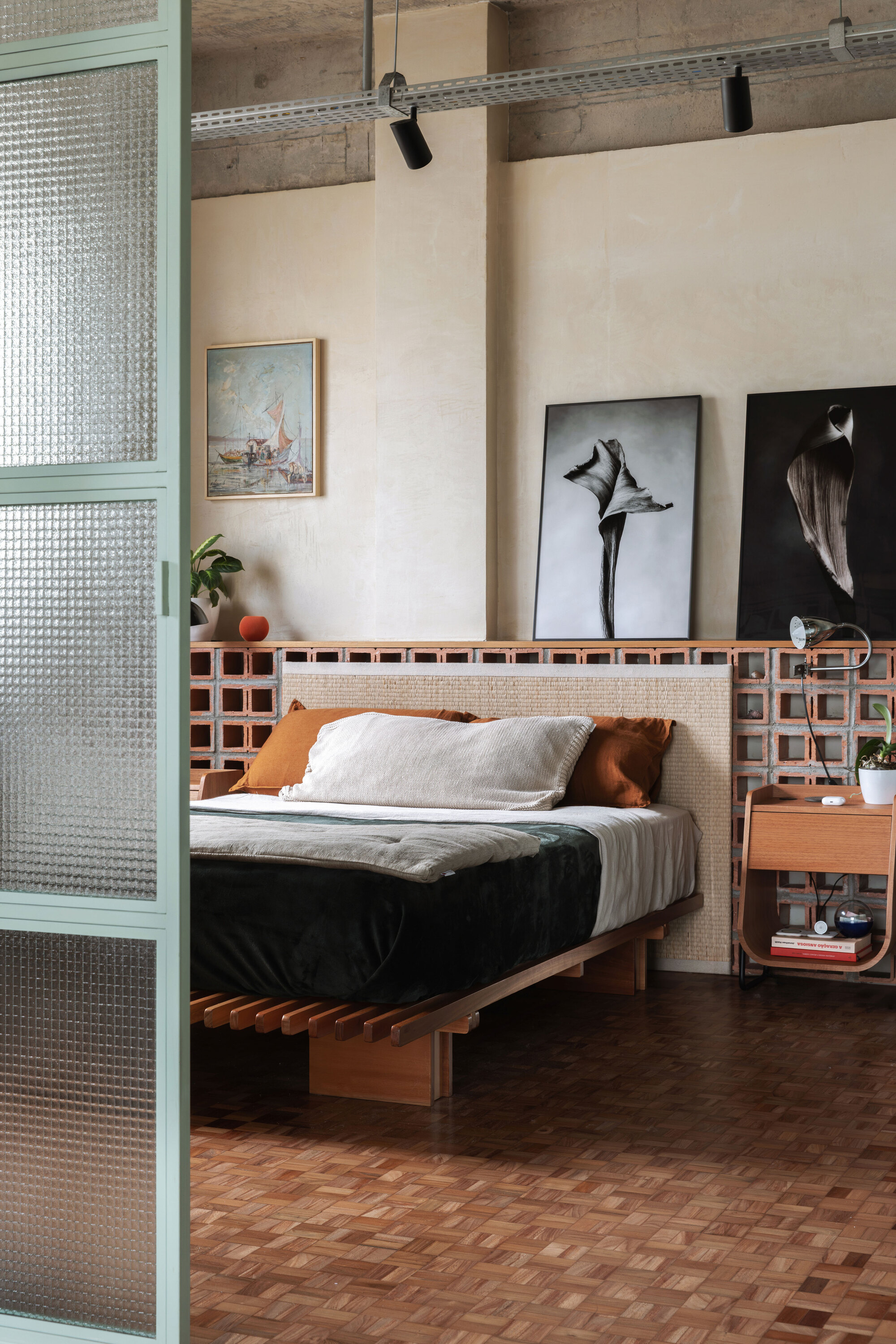 Bedroom with a slatted timber bed against a cobogó block half wall, with large black and white botanical prints and track lighting overhead