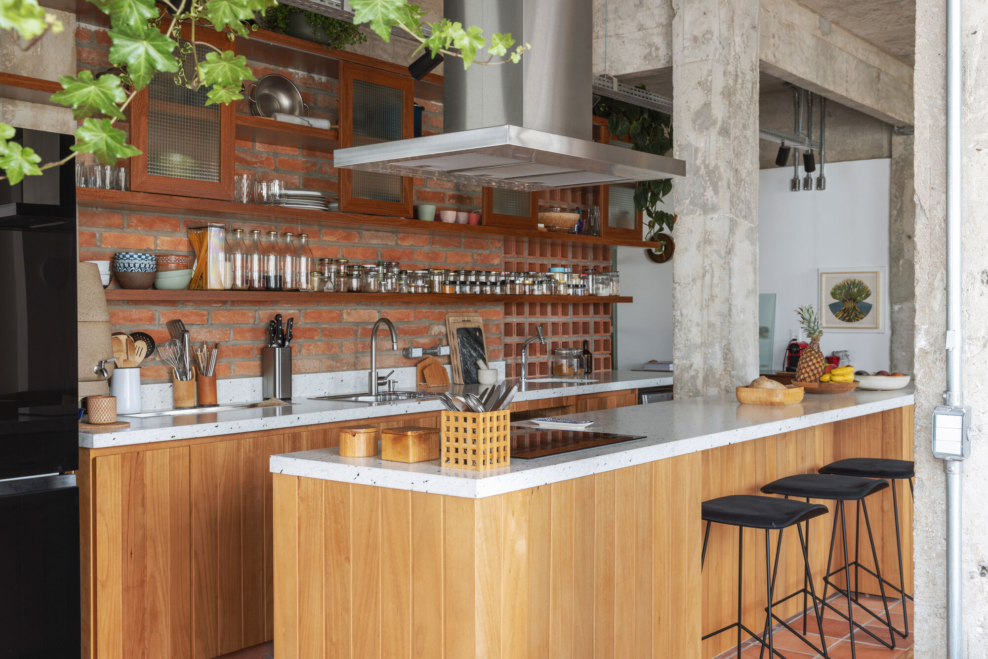 Kitchen with exposed brick backsplash, timber-clad island with terrazzo counter, black stools and a steel extractor hood beside raw concrete columns