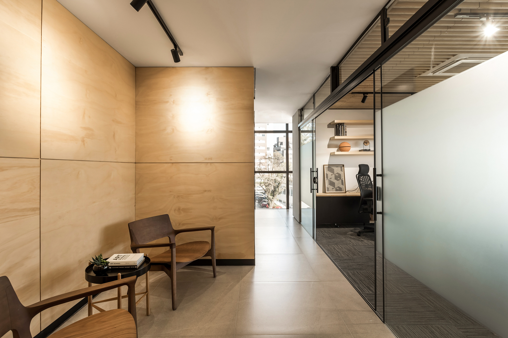 Corridor with plywood wall panels, two wooden armchairs and a frosted glass partition opening onto a private office, lit by a window at the end