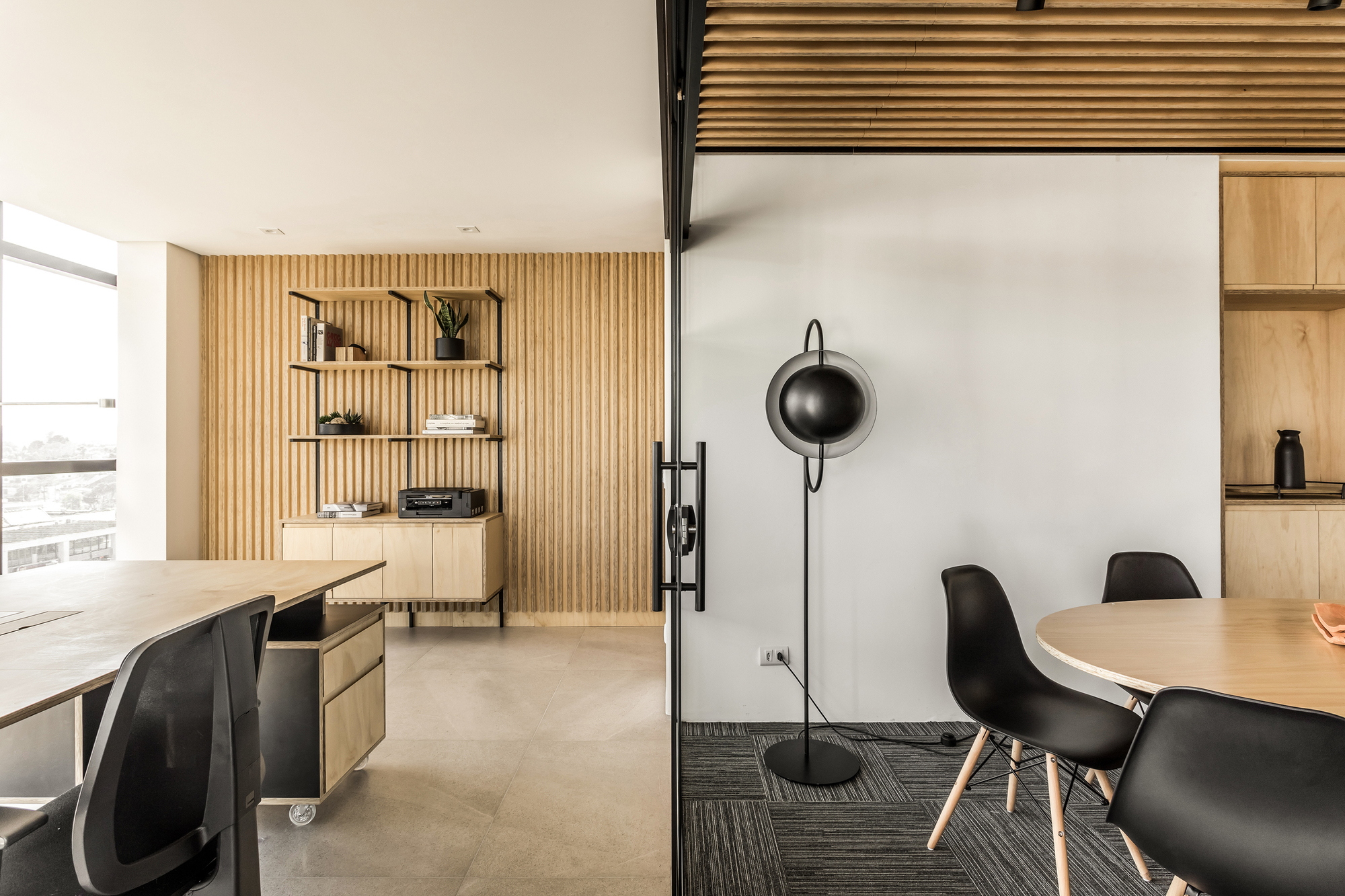 Vertical timber slat wall with open shelving behind a desk, seen through a glass door past an oval table and black chairs