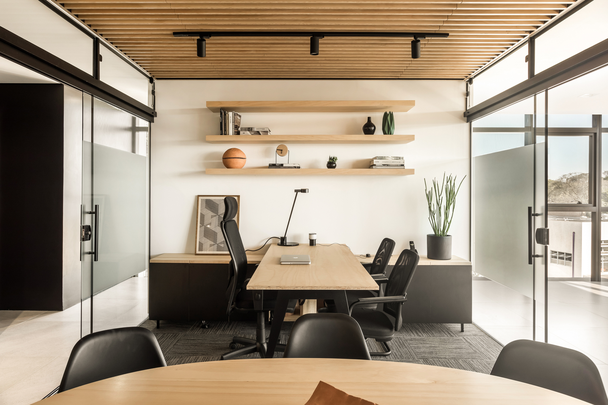 Private office with floating oak shelves, a timber desk and black mesh chairs, framed by glass walls and a slatted ceiling in daylight