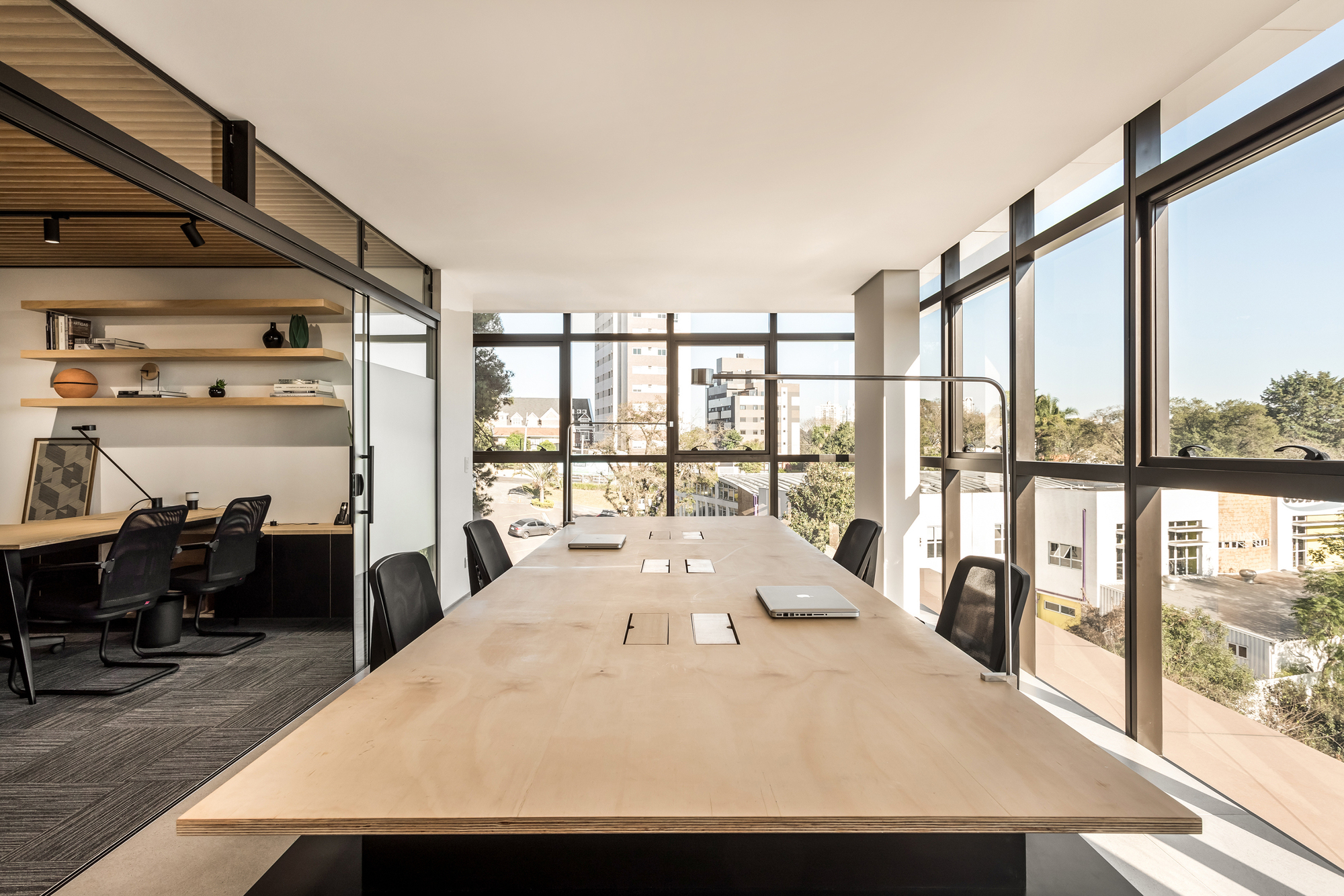Long plywood conference table facing corner windows with treetop views, beside a glass-walled office, under clear afternoon sky