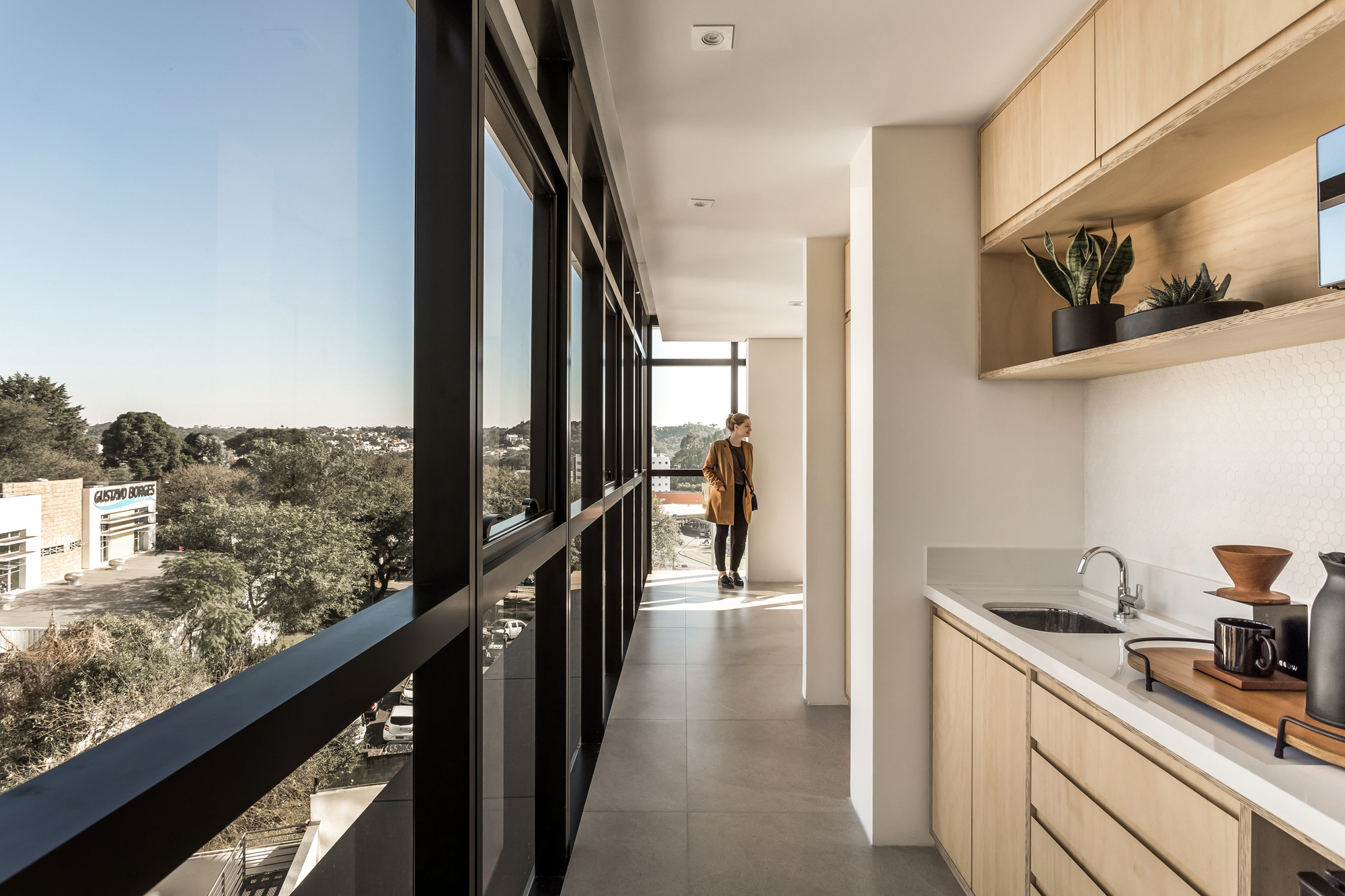 Narrow pantry corridor with a plywood kitchenette and planted shelf, a woman in a mustard coat standing beside the floor-to-ceiling window wall