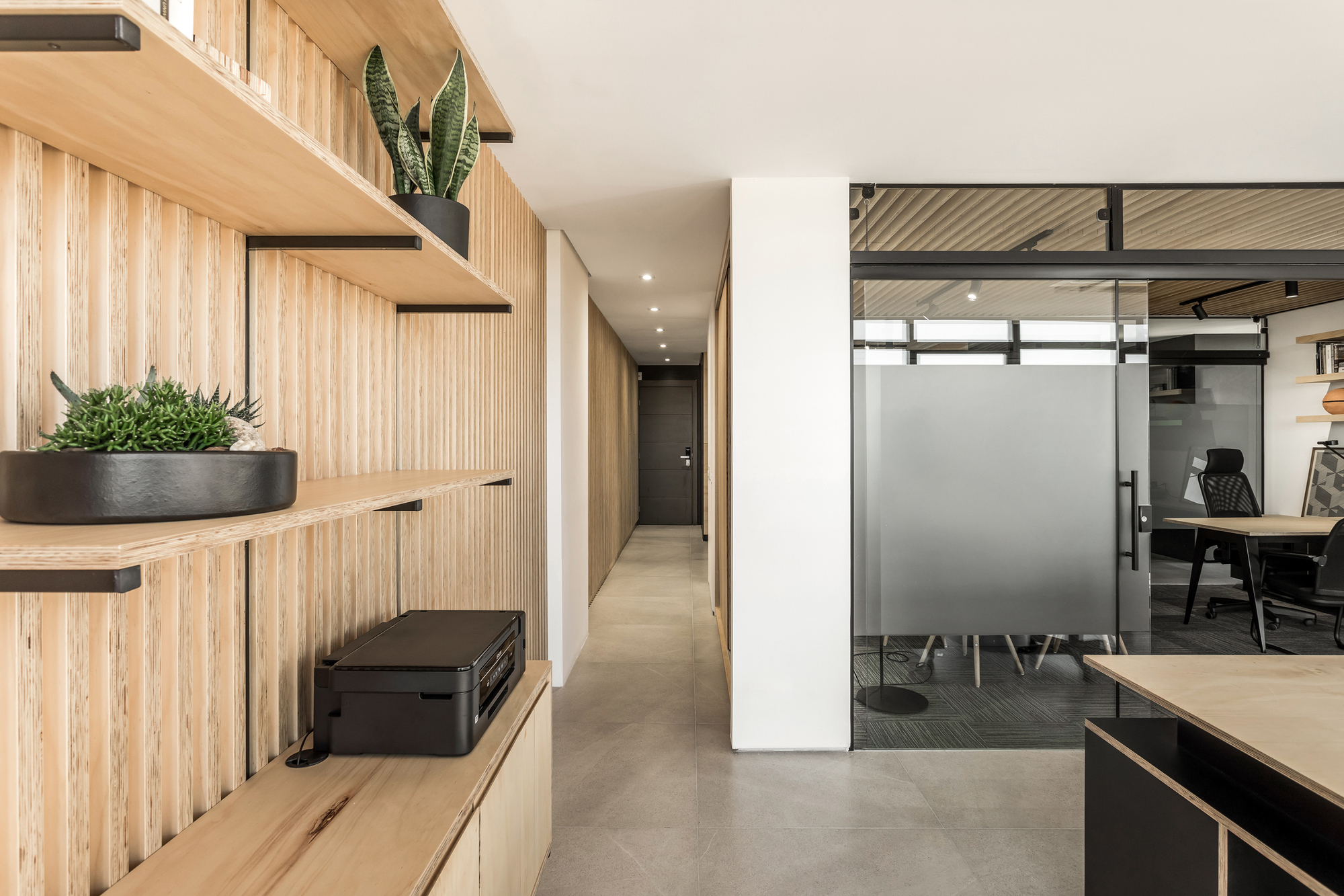 Corridor lined with vertical timber slats and floating shelves holding potted plants and a printer, beside a frosted glass meeting room