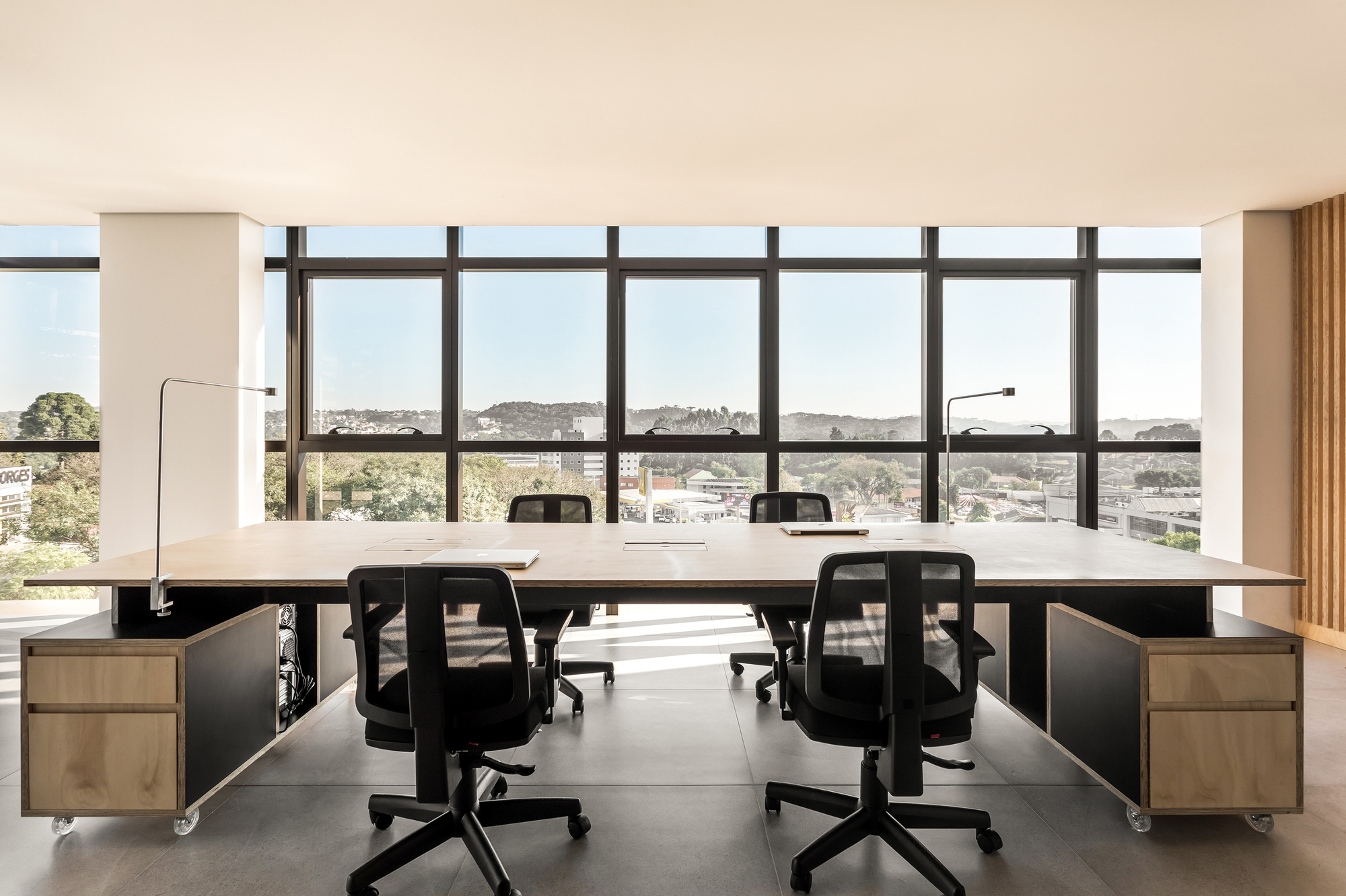 Shared plywood work table with rolling drawer units facing a glazed wall and hillside view in bright daylight