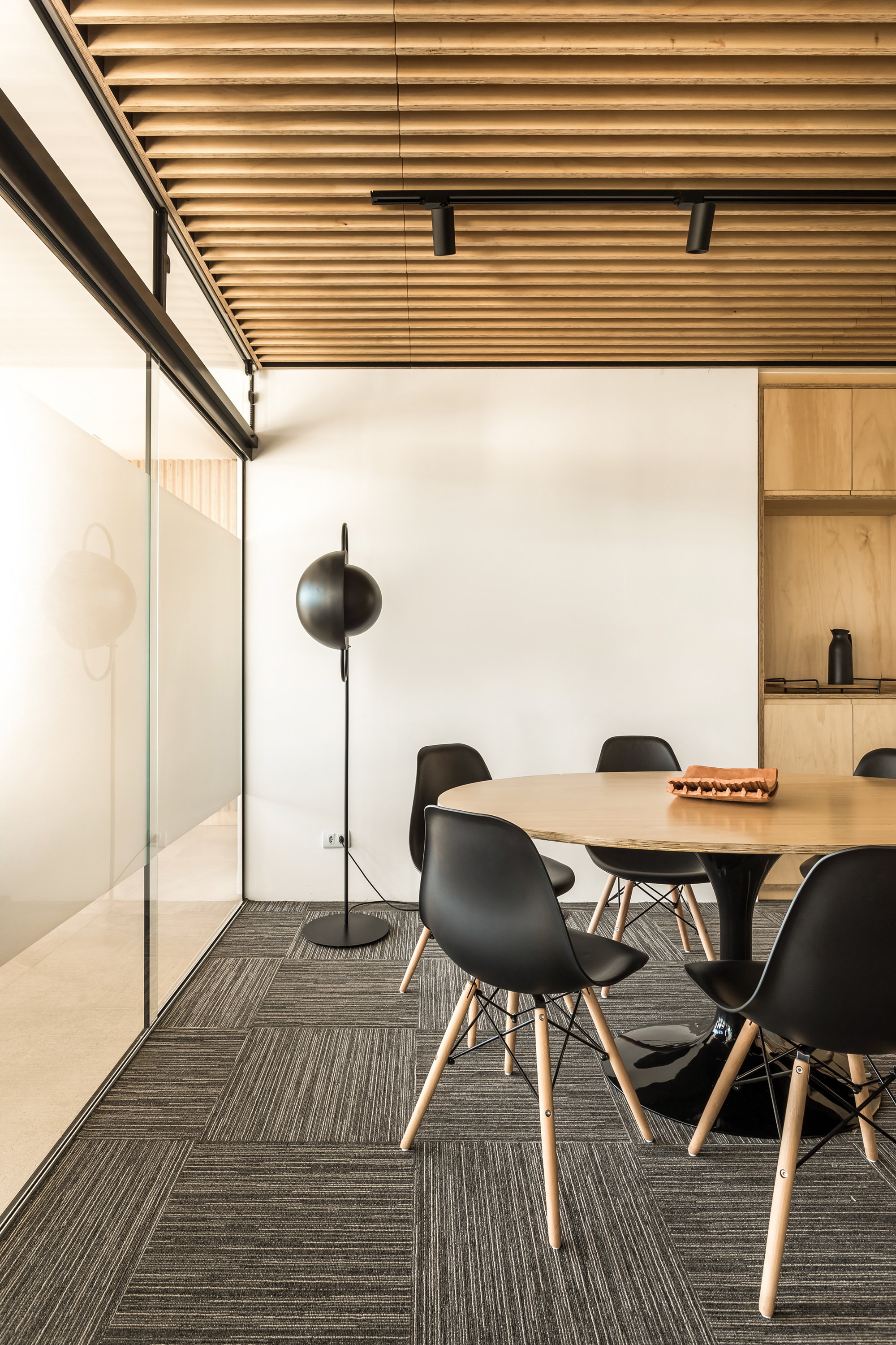 Meeting corner with round table, black shell chairs, slatted timber ceiling and a globe floor lamp beside frosted glazing