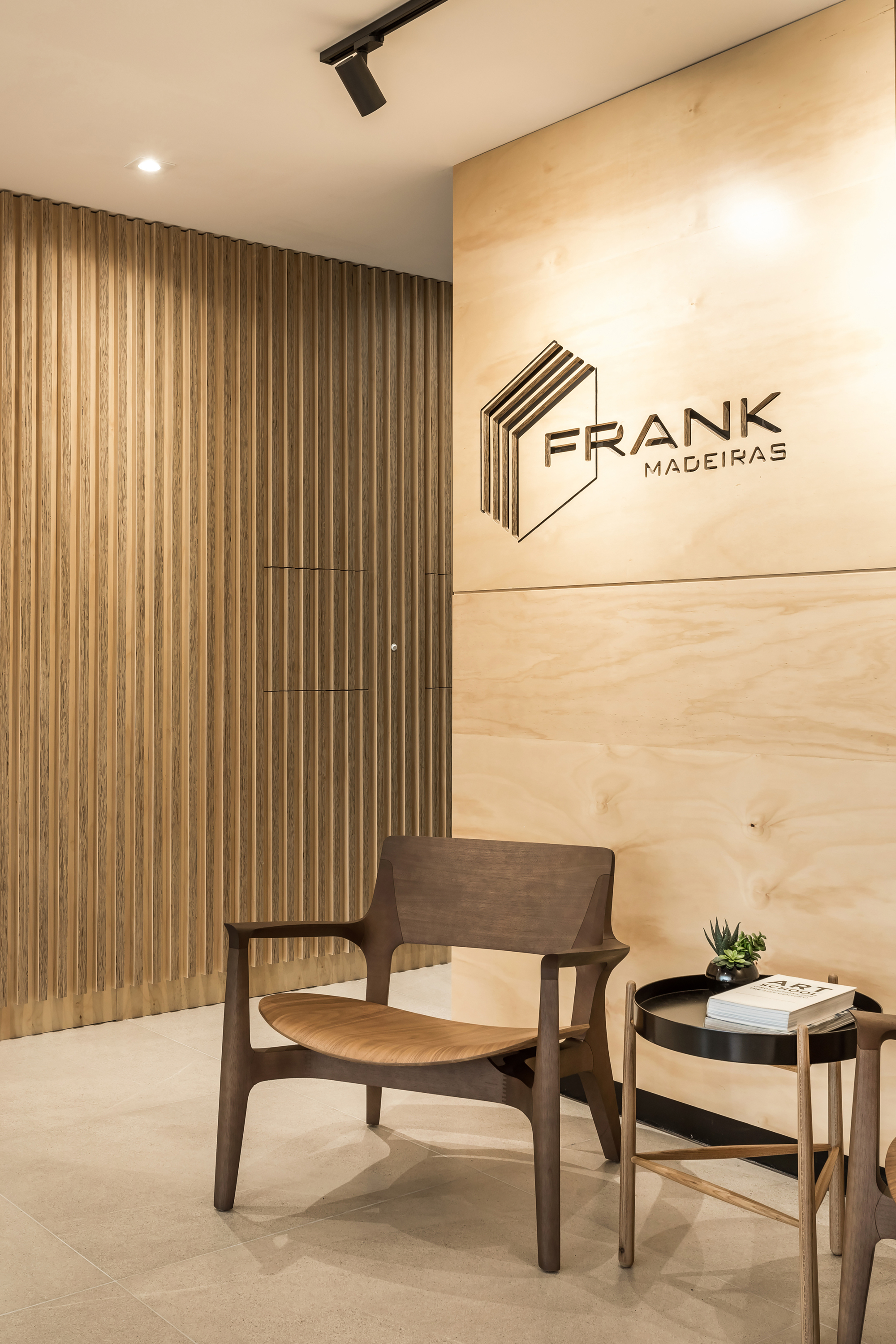 Reception corner with a wooden lounge chair and side table in front of a plywood logo wall and fluted timber panelling