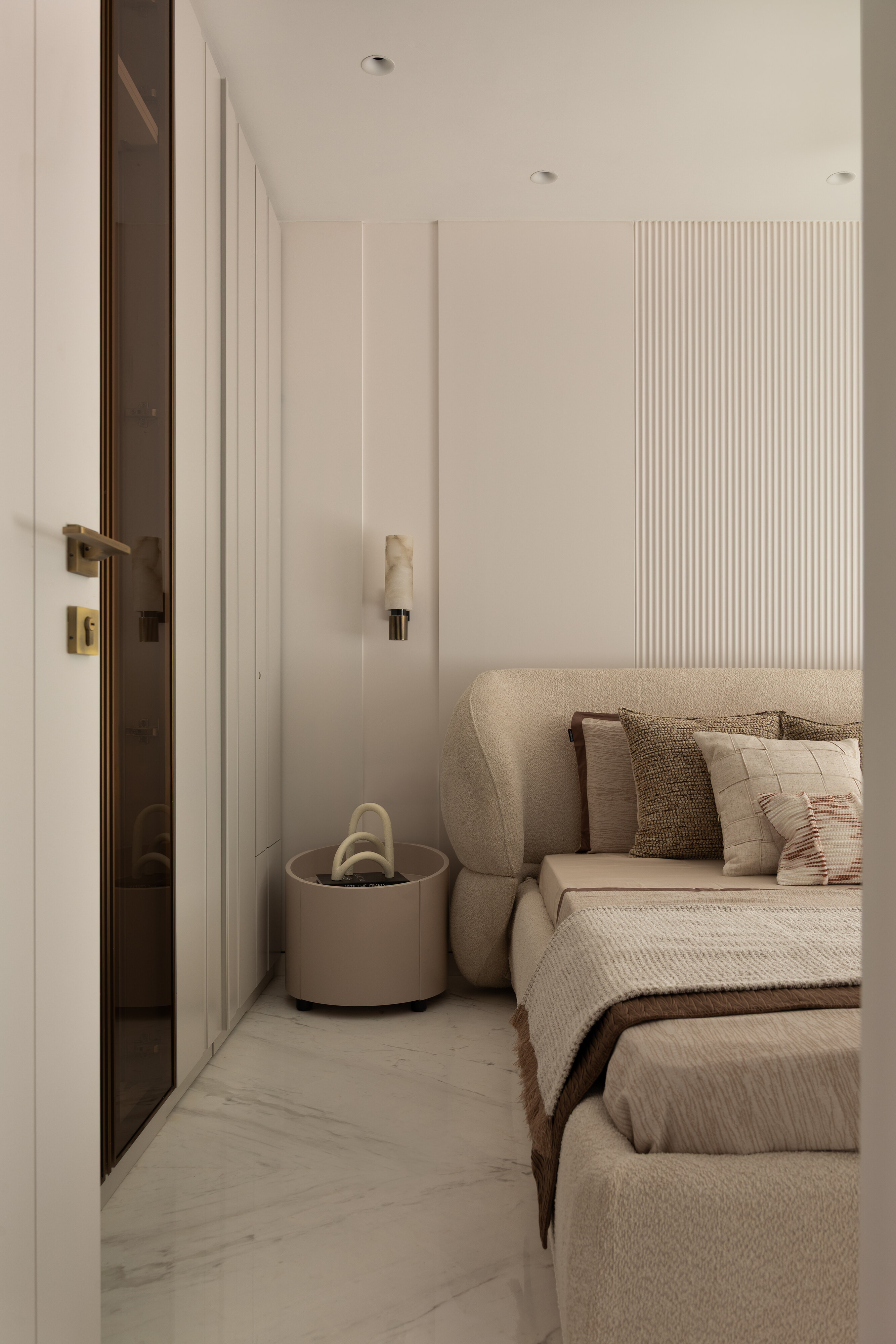 A side view of the bouclé bed alongside a tinted brass-framed glass door and an alabaster wall sconce.