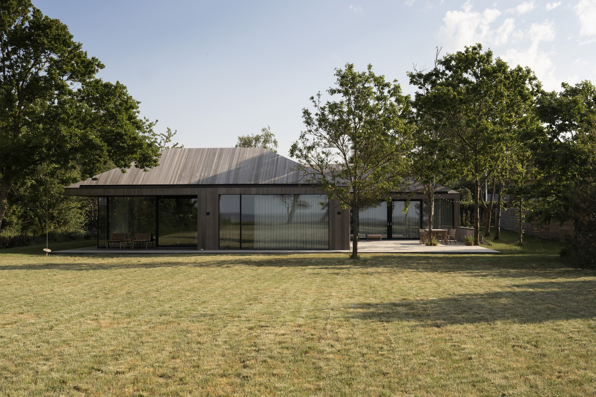 Long view of the single-story residence with corrugated metal roof and glass walls set among trees in dry grass