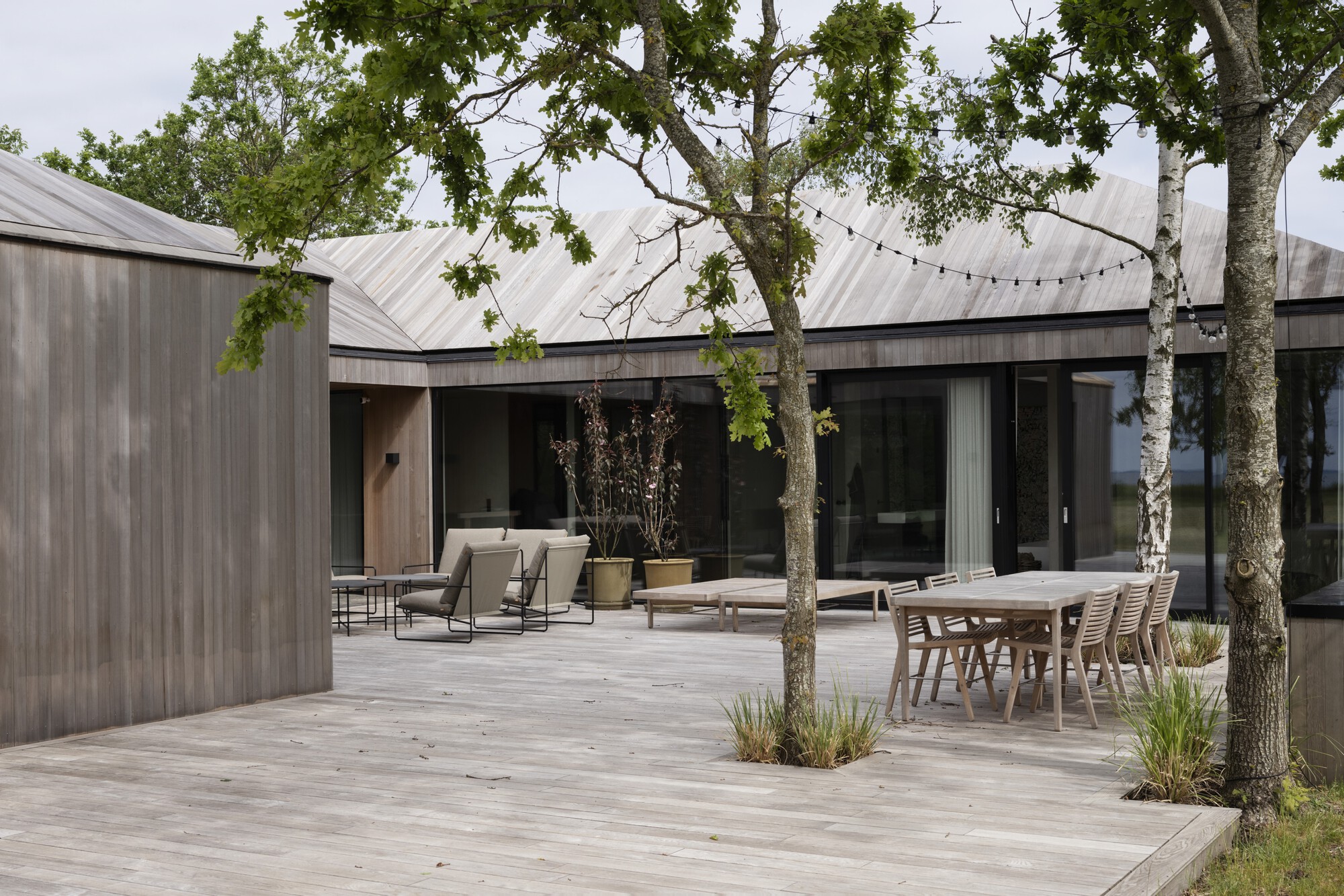 Timber deck courtyard with outdoor dining furniture and lounge chairs beneath overhanging tree branches and string lights