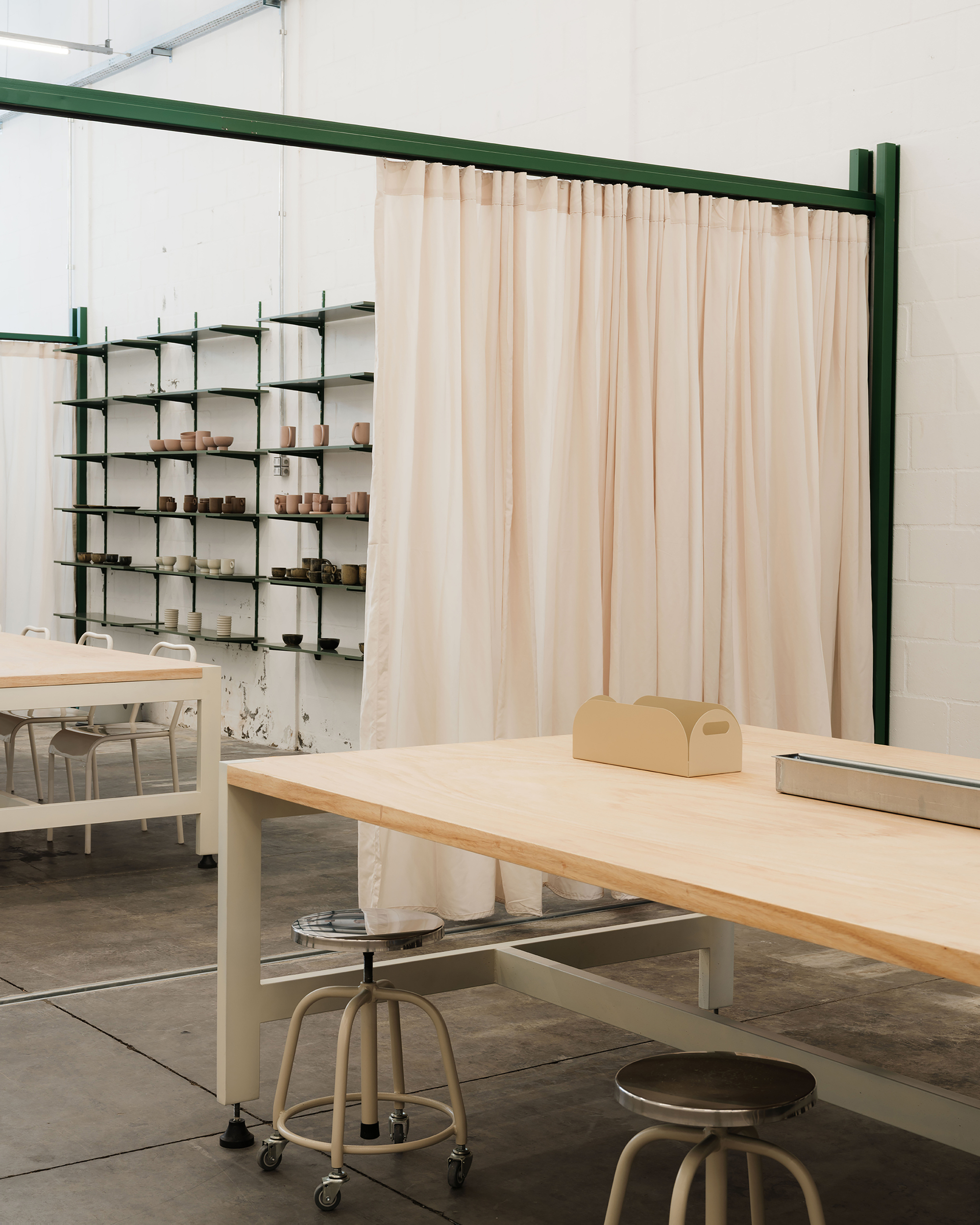 Plywood worktable with adjustable metal stools and shelving unit displaying ceramic objects