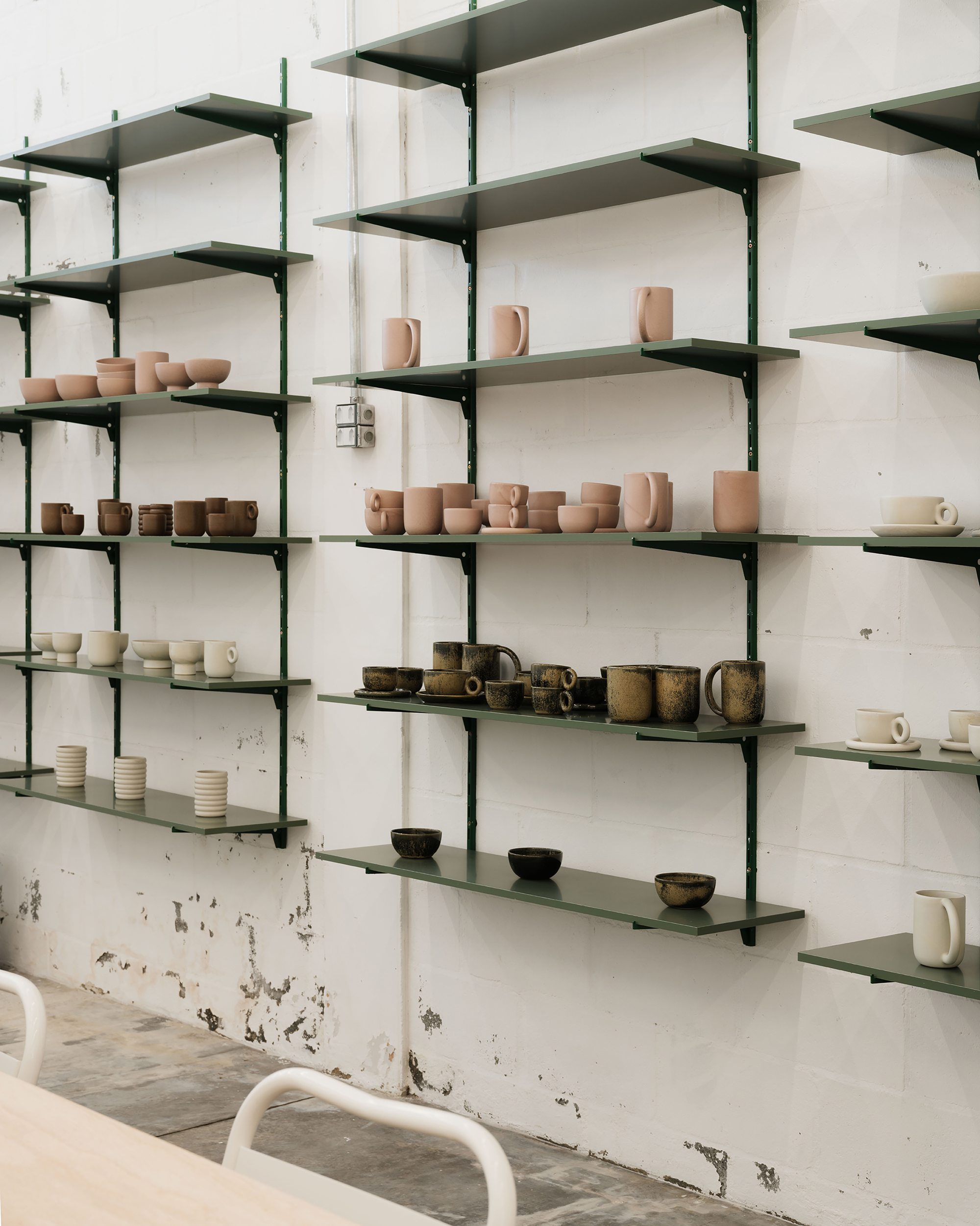 Green steel shelving system mounted on white tile wall displaying handmade ceramic vessels and cups