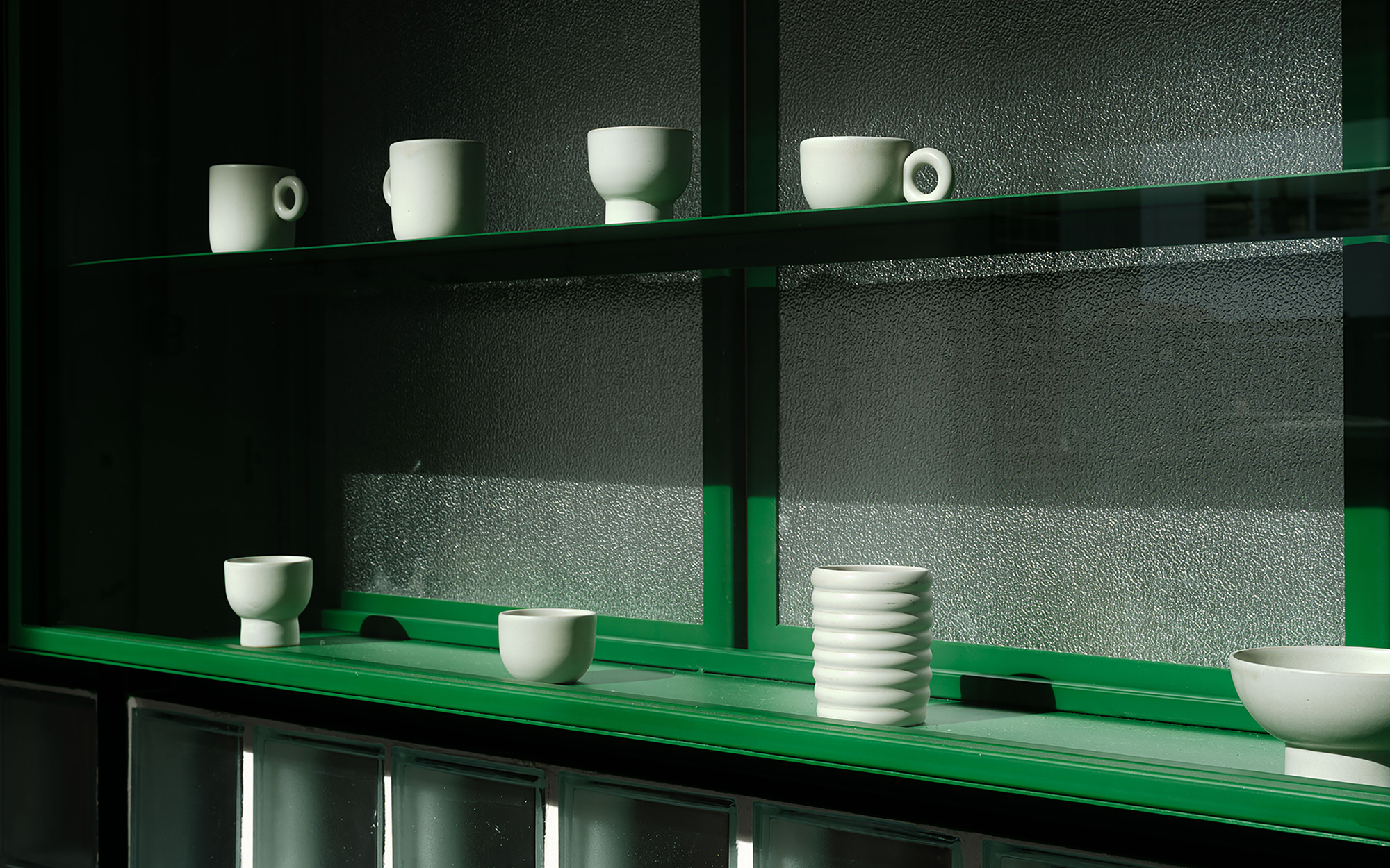Close-up of green powder-coated steel shelves holding white ceramic cups against textured glass panels