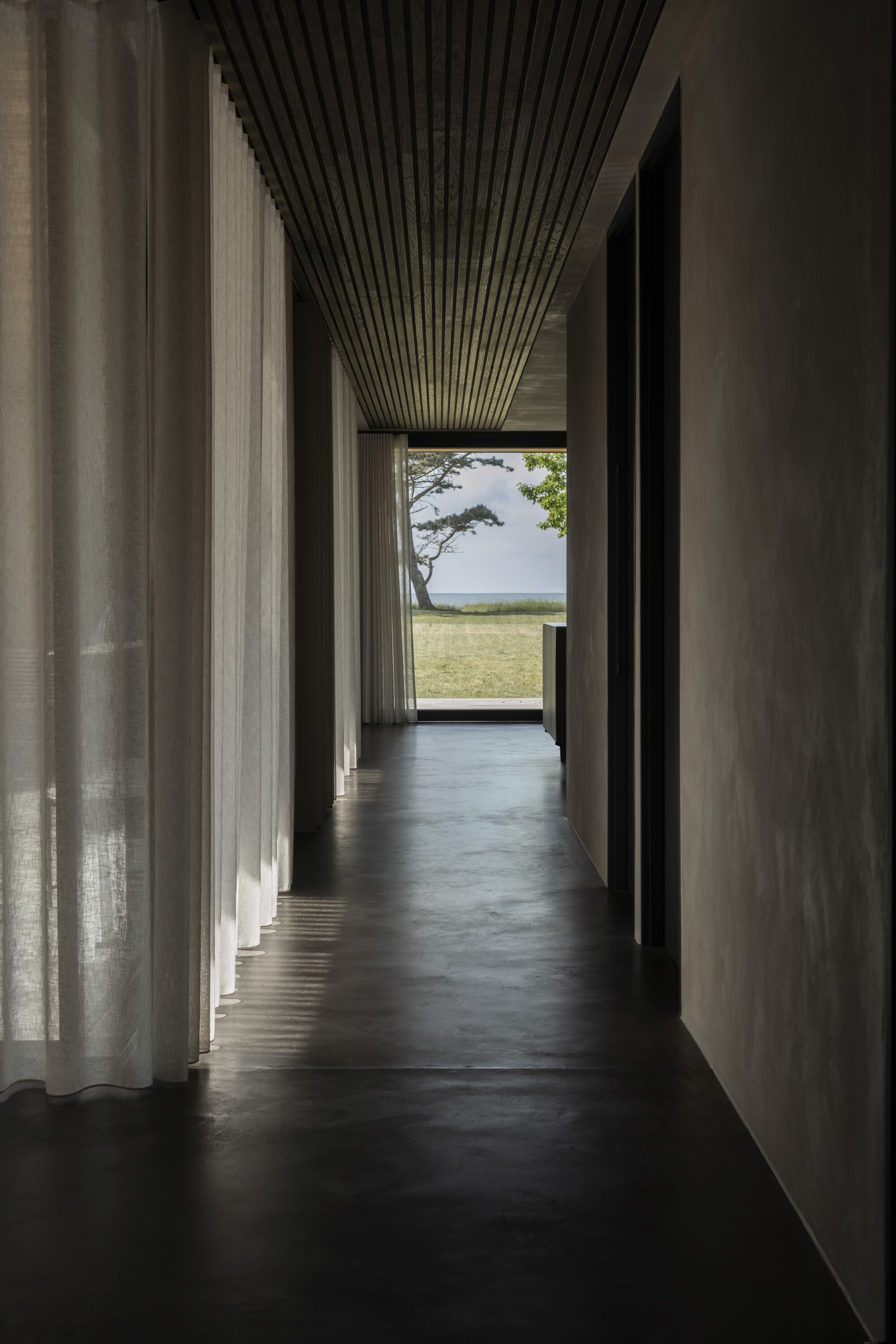 Dark polished concrete corridor with ribbed ceiling and sheer curtains framing a view to open fields