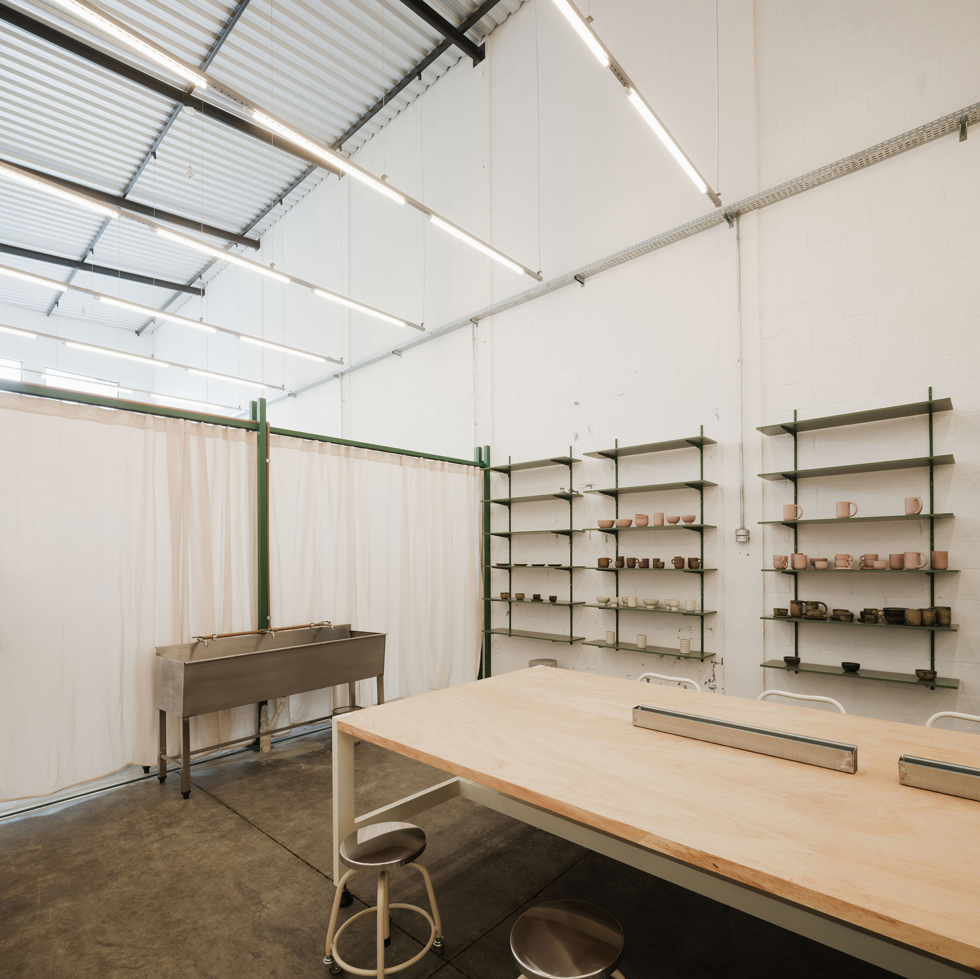 Workshop interior with plywood work table, green shelving, sheer curtain partition, and corrugated metal ceiling