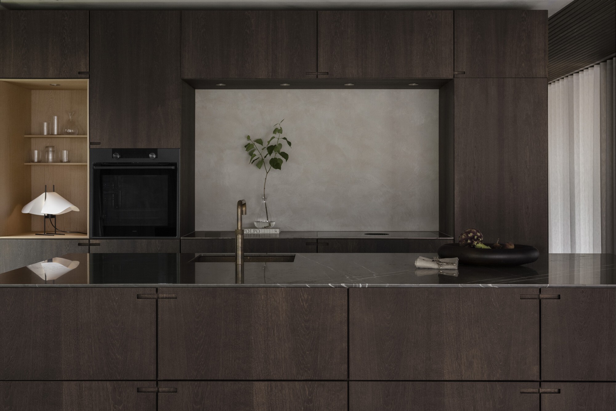 Kitchen island in dark timber with integrated sink facing a built-in oven and shelving niche