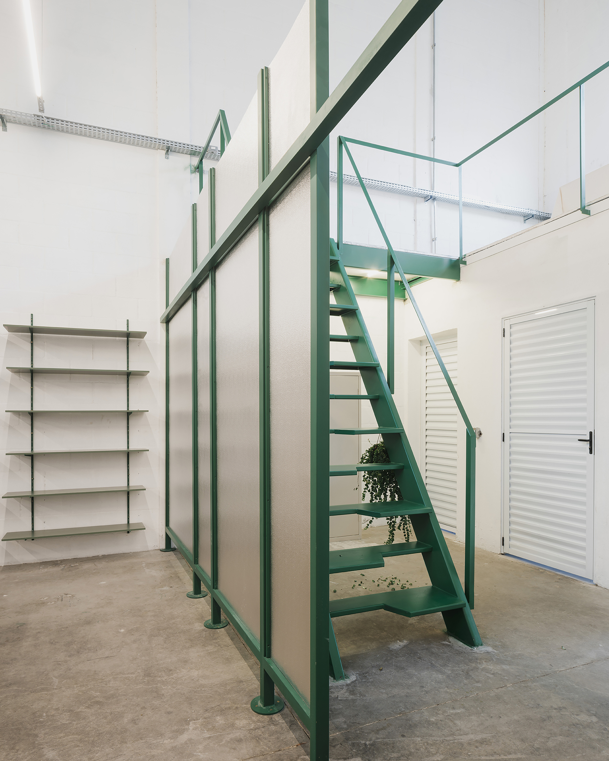 Double-height space with green steel staircase, glass railing above, and wall-mounted shelving under polished concrete floor