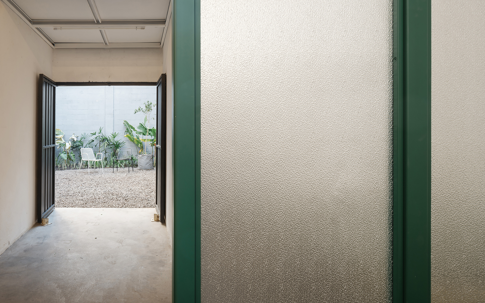 View through hallway with textured white wall trimmed in green steel toward courtyard with gravel and plants