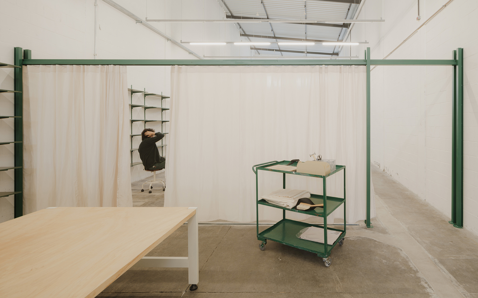 Workspace with green steel frame, fabric curtains, plywood table and rolling cart under exposed industrial ceiling