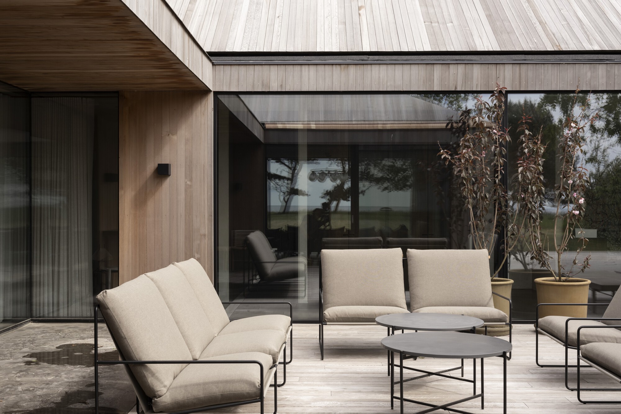 Terrace with timber-clad volumes and floor-to-ceiling glazing framed by upholstered lounge furniture and potted plants