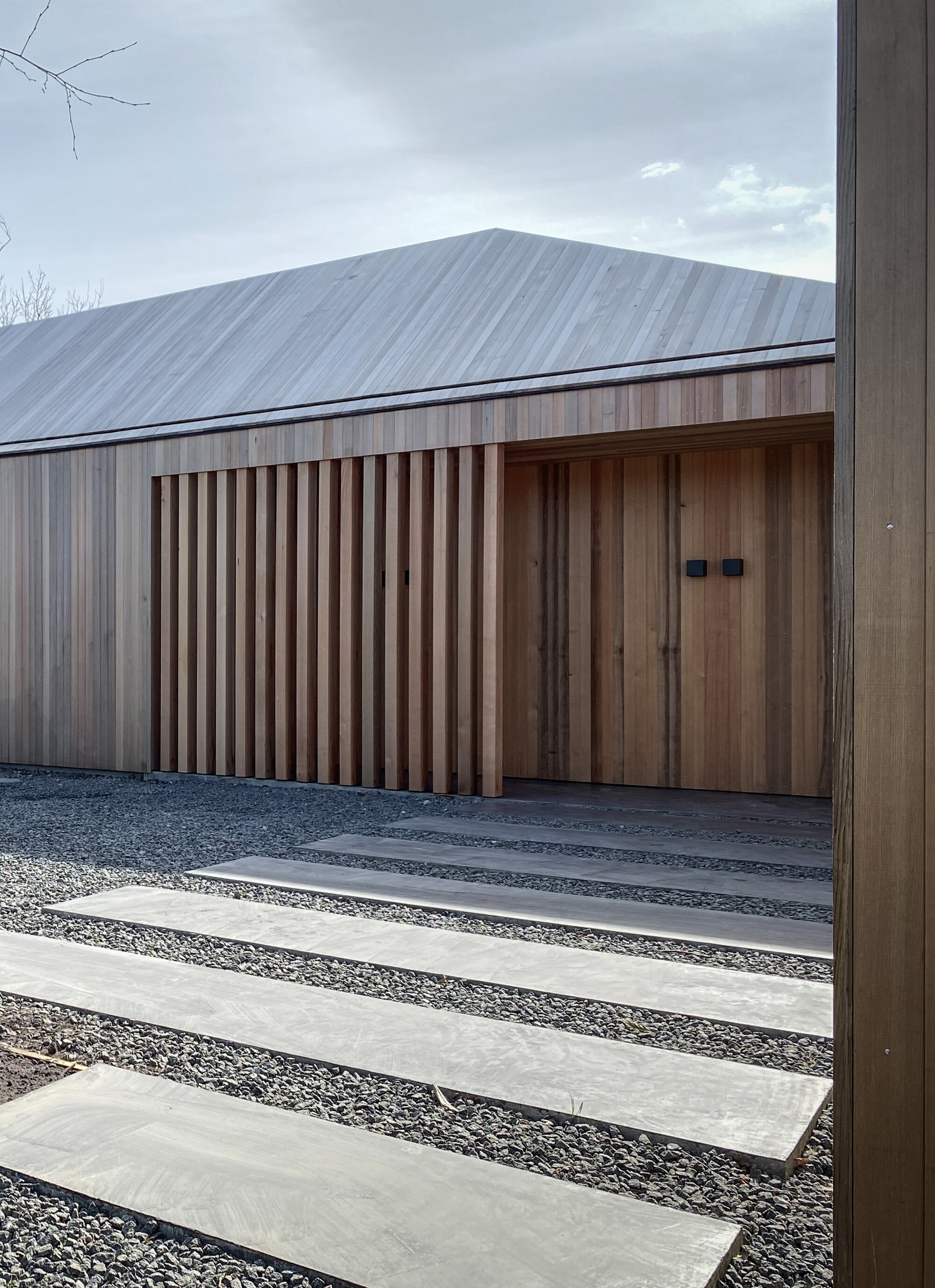 Timber-slatted facade with concealed door alongside concrete pavers set in gravel under overcast skies