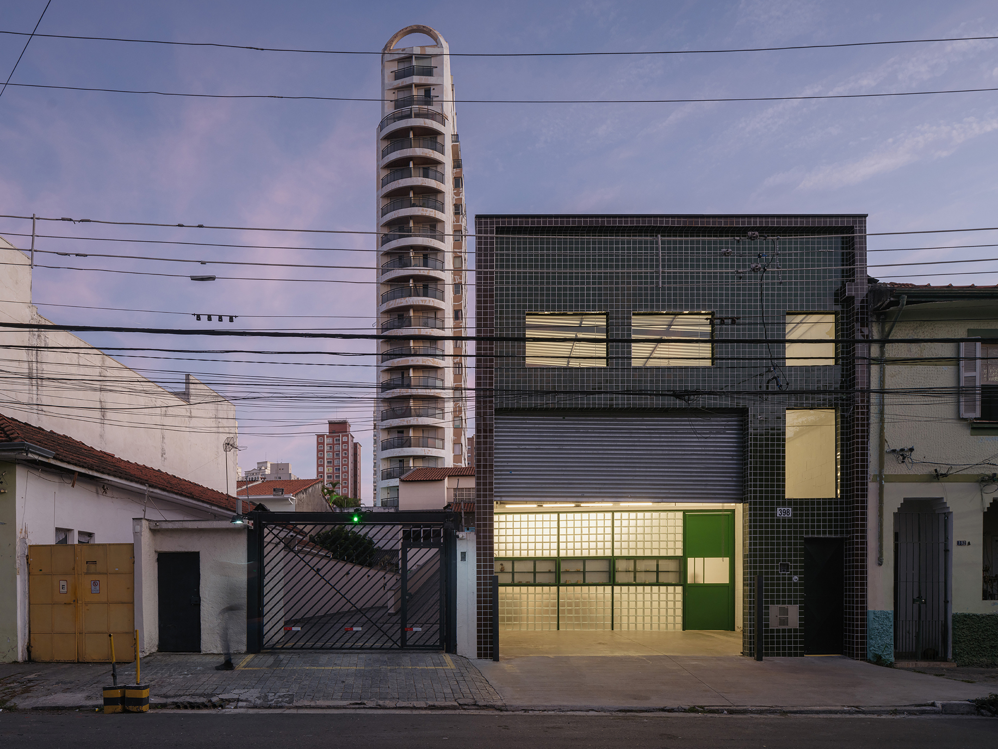 Evening view of illuminated facade with glass block and green tile next to neighboring tower under purple sky