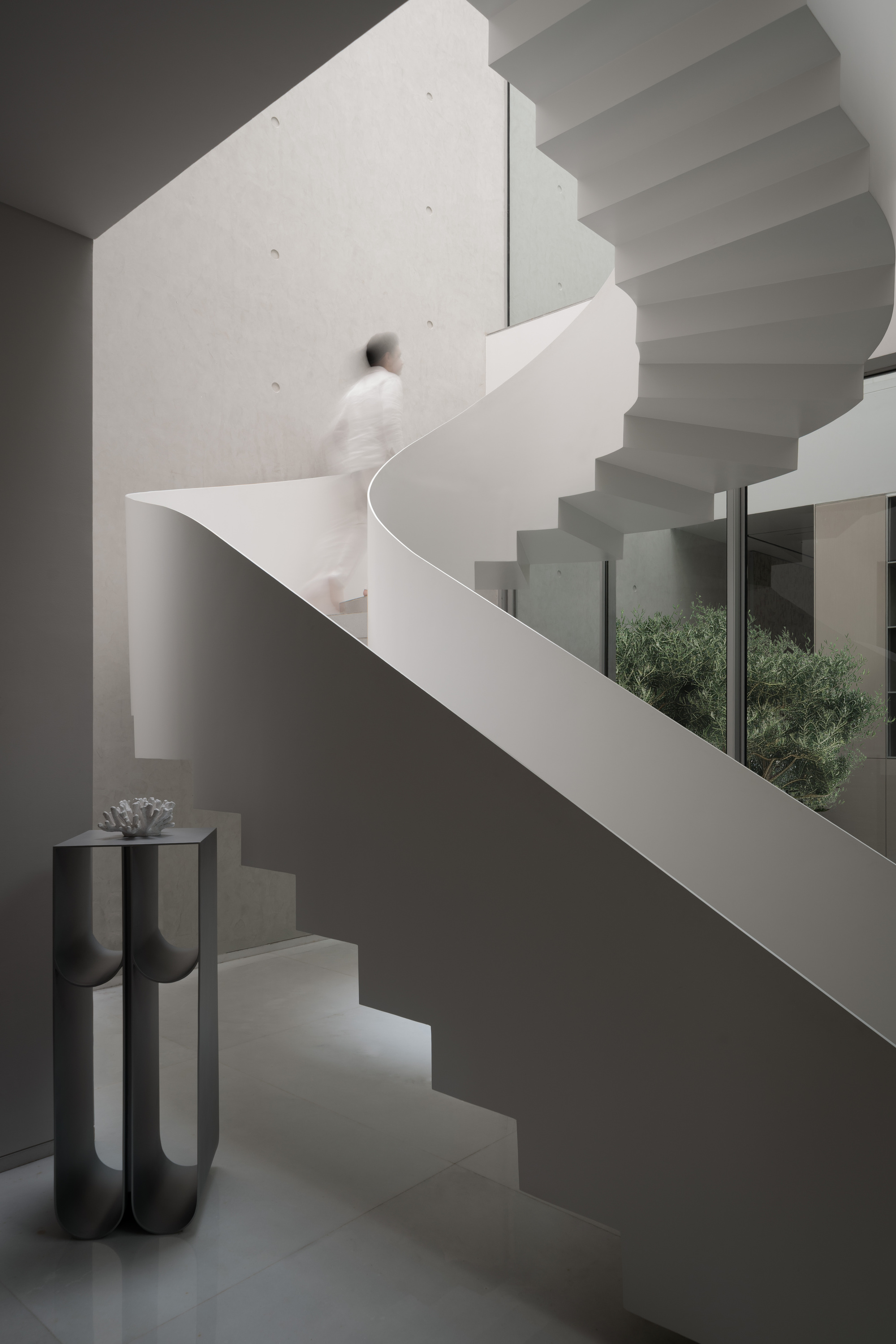 White sculptural staircase with a person ascending past a black pedestal and courtyard view