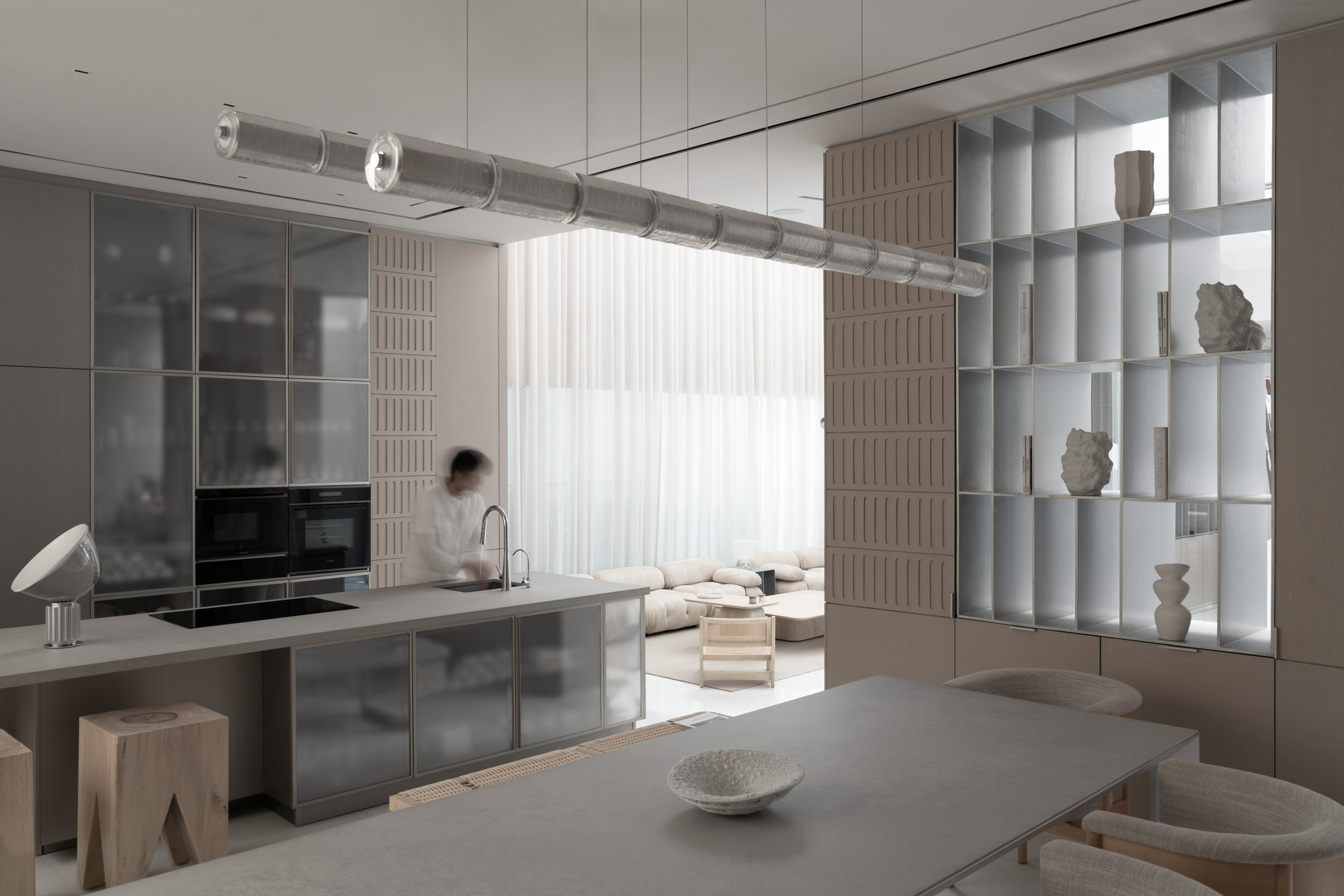 Open kitchen with reflective stainless steel island beneath a tubular glass pendant light and gridded display wall