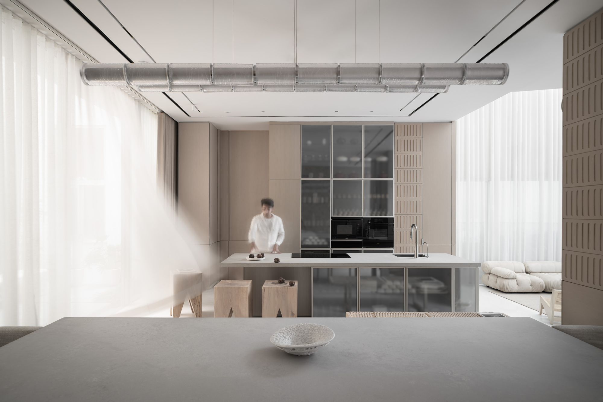Open kitchen with exposed ducting overhead and a person standing at the island beneath sheer curtains